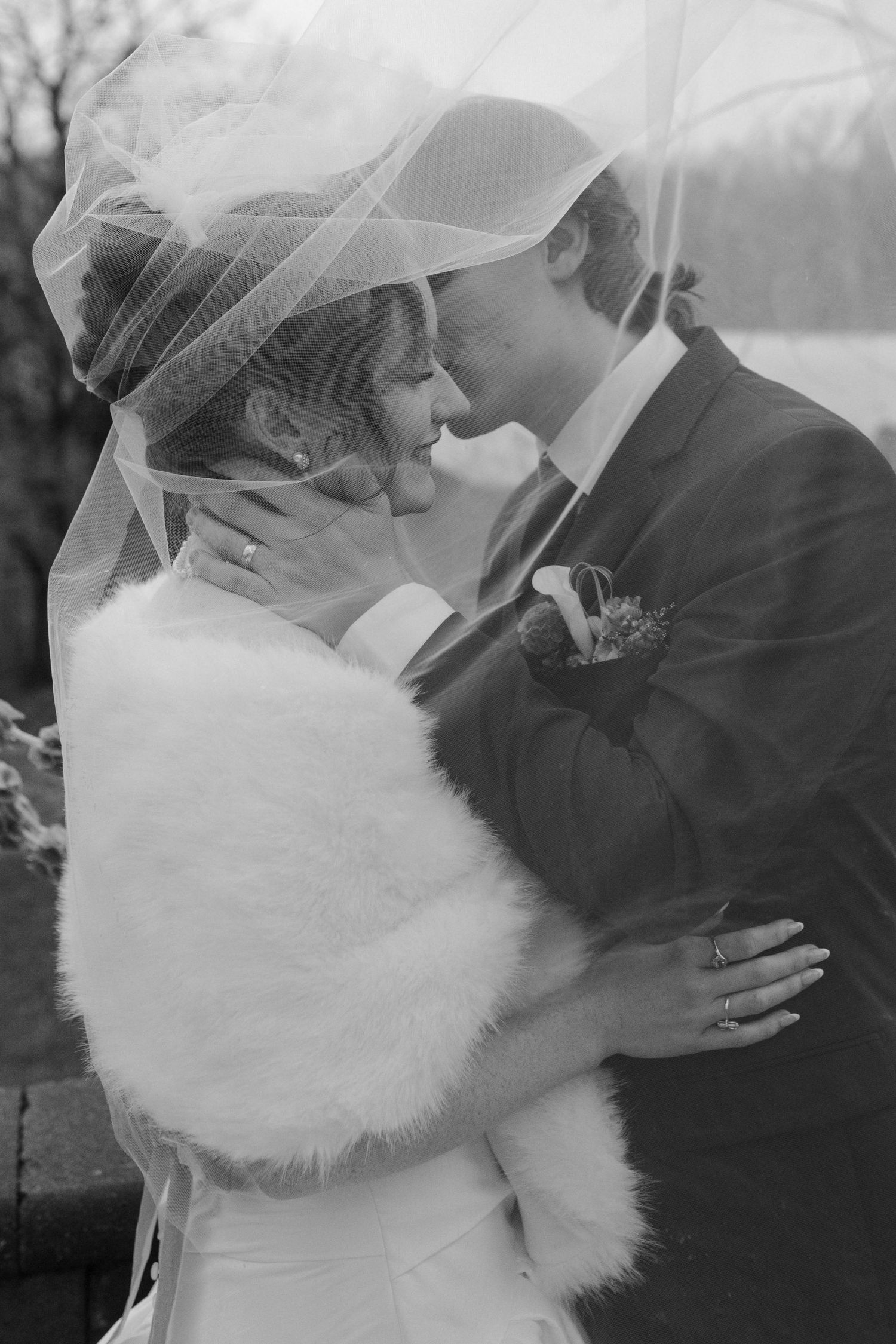 Black and white photo of a bride and groom sharing a kiss under a veil outdoors in winter.
