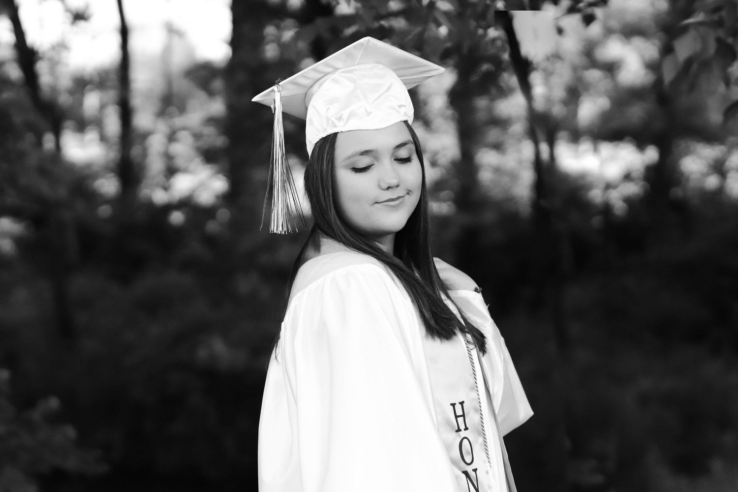 Black and white photo of a young woman in white graduation cap and gown smiling outdoors.