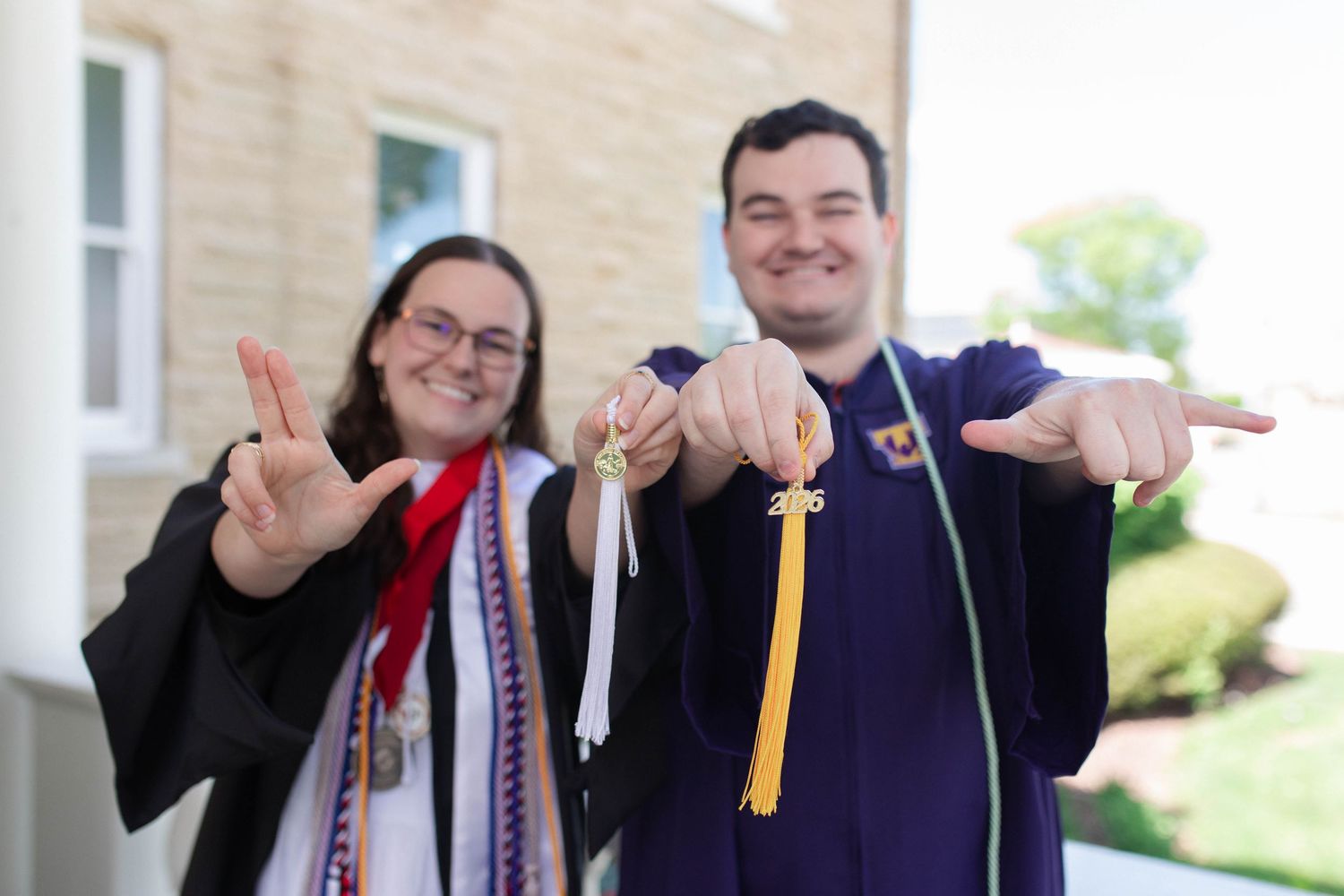 Two graduates in caps and gowns with honor cords hold up keys and peace signs outside a brick building.