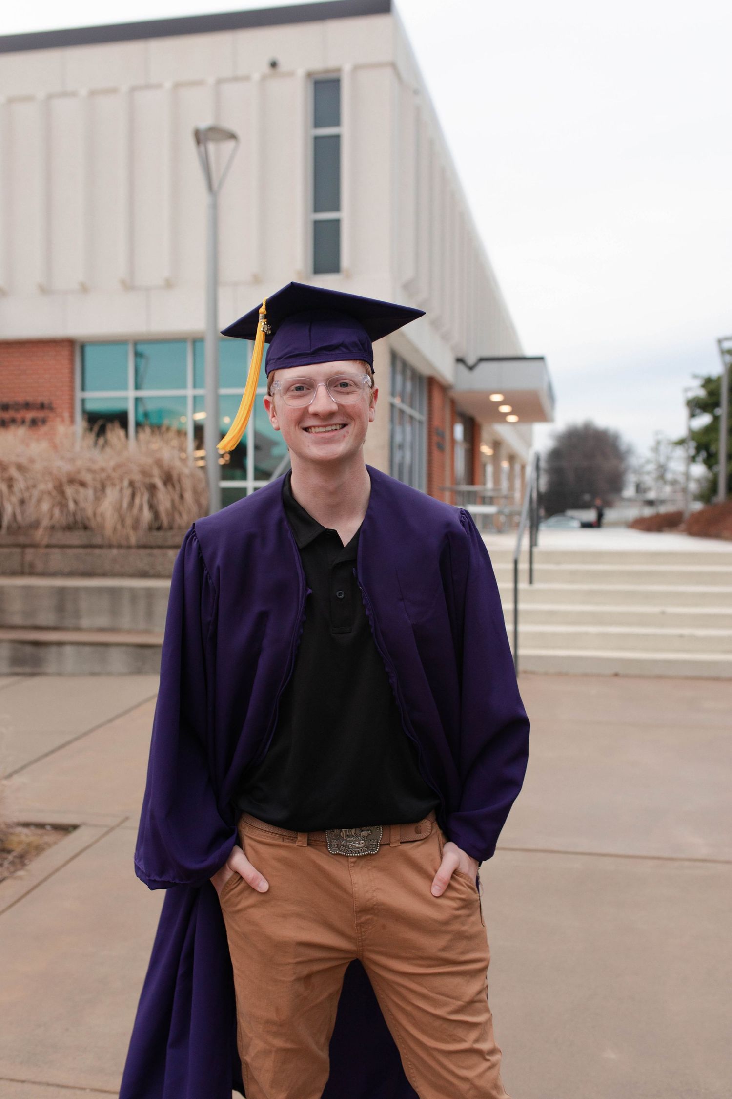 Young man smiling in purple graduation cap and gown standing outside a school building on graduation day.