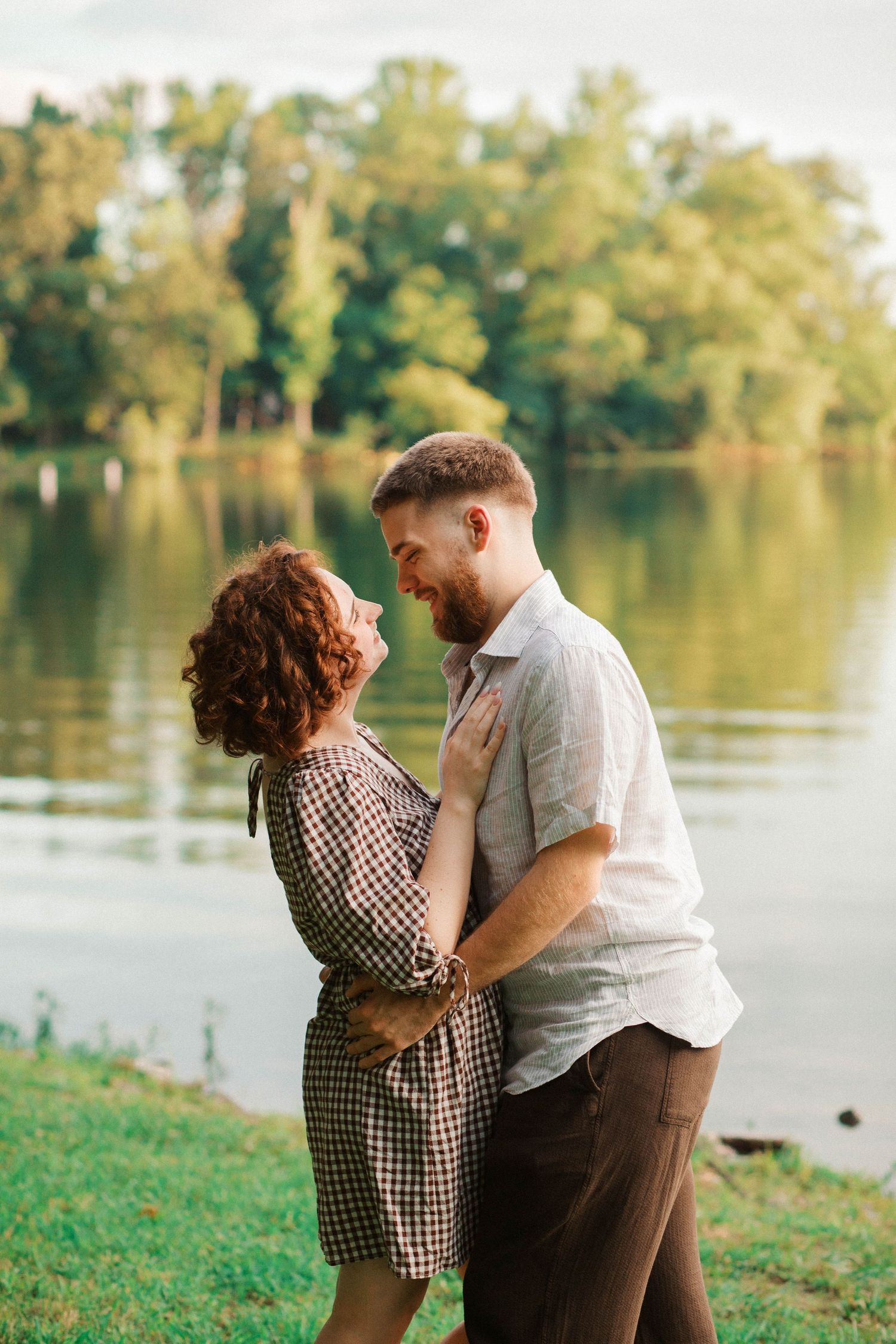 Romantic couple embracing and smiling by a serene lake surrounded by lush green trees on a warm day.