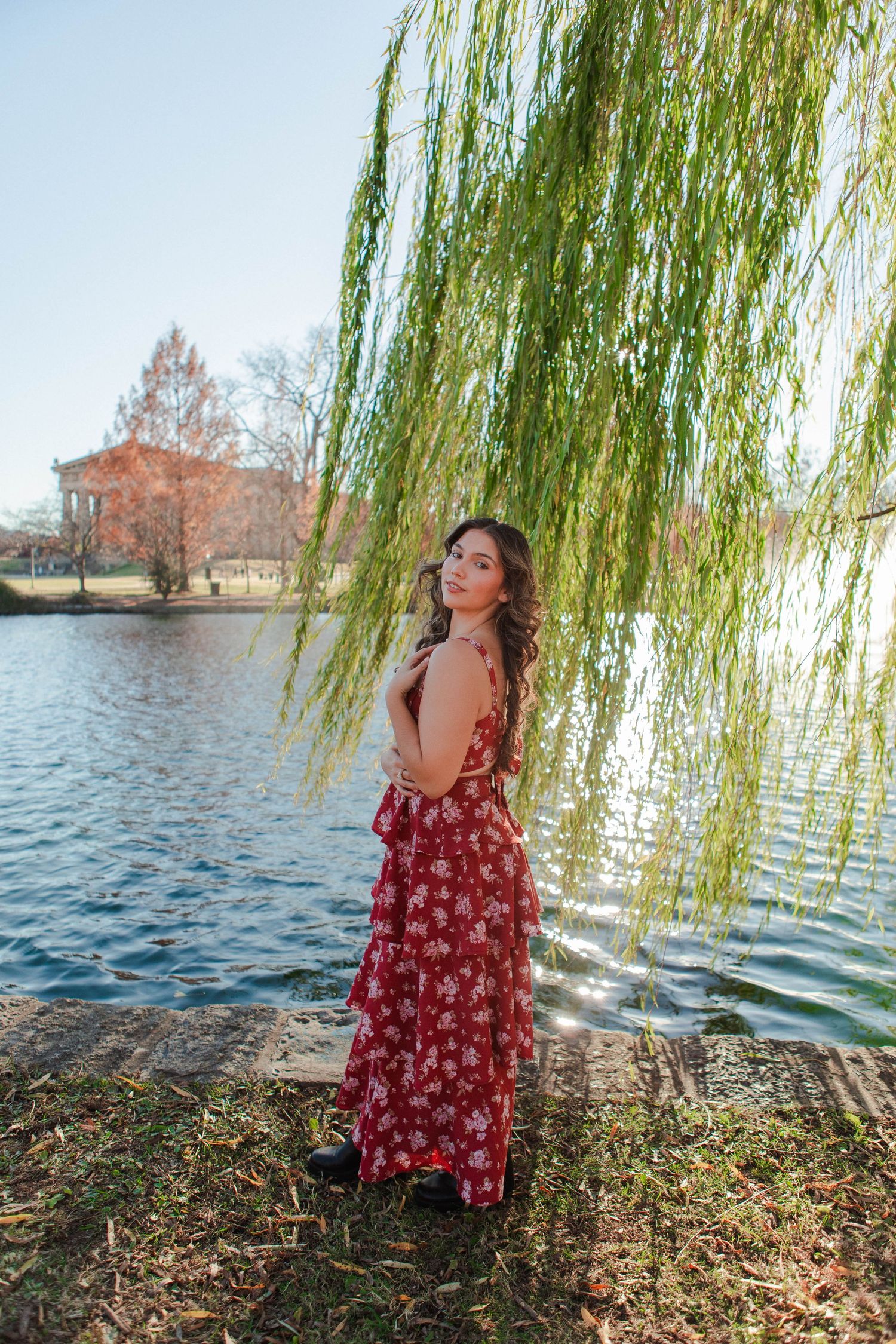 Young woman in red floral maxi dress standing by a serene lake under a weeping willow tree on a sunny day.