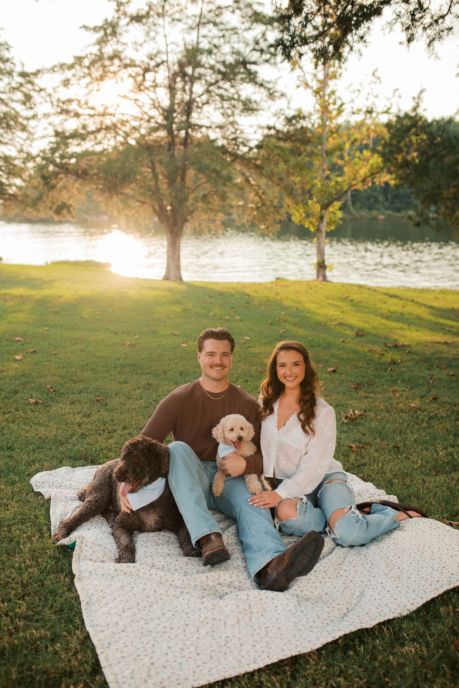 Couple sitting on blanket by lake at sunset with two dogs, one brown and one cream colored.