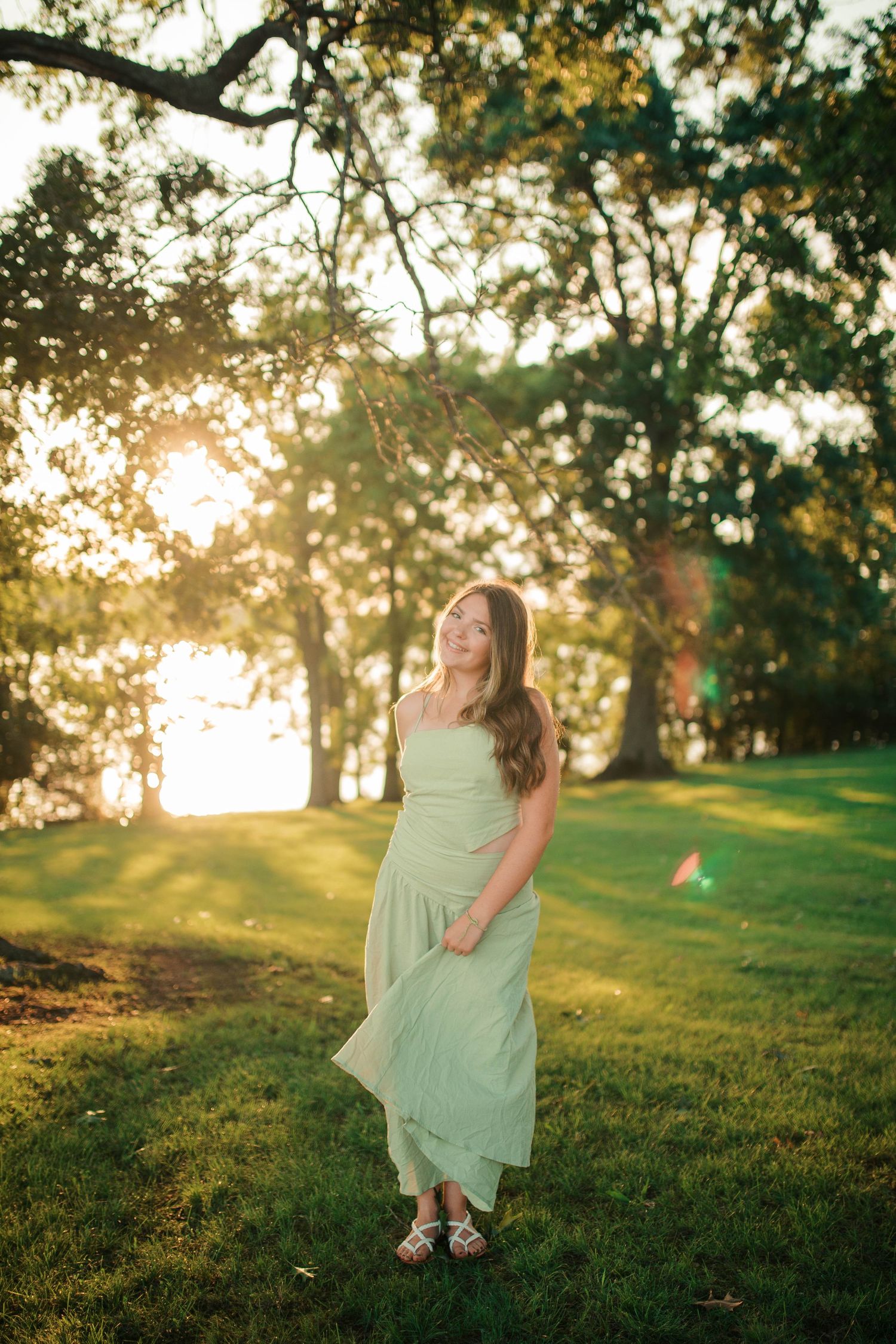 Young woman in mint green dress standing on lush grass at golden hour sunset in a park.
