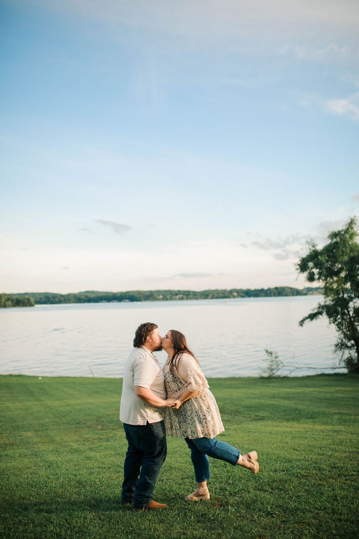 Couple sharing a romantic kiss on green grass by a serene lake at sunset during an outdoor engagement session.
