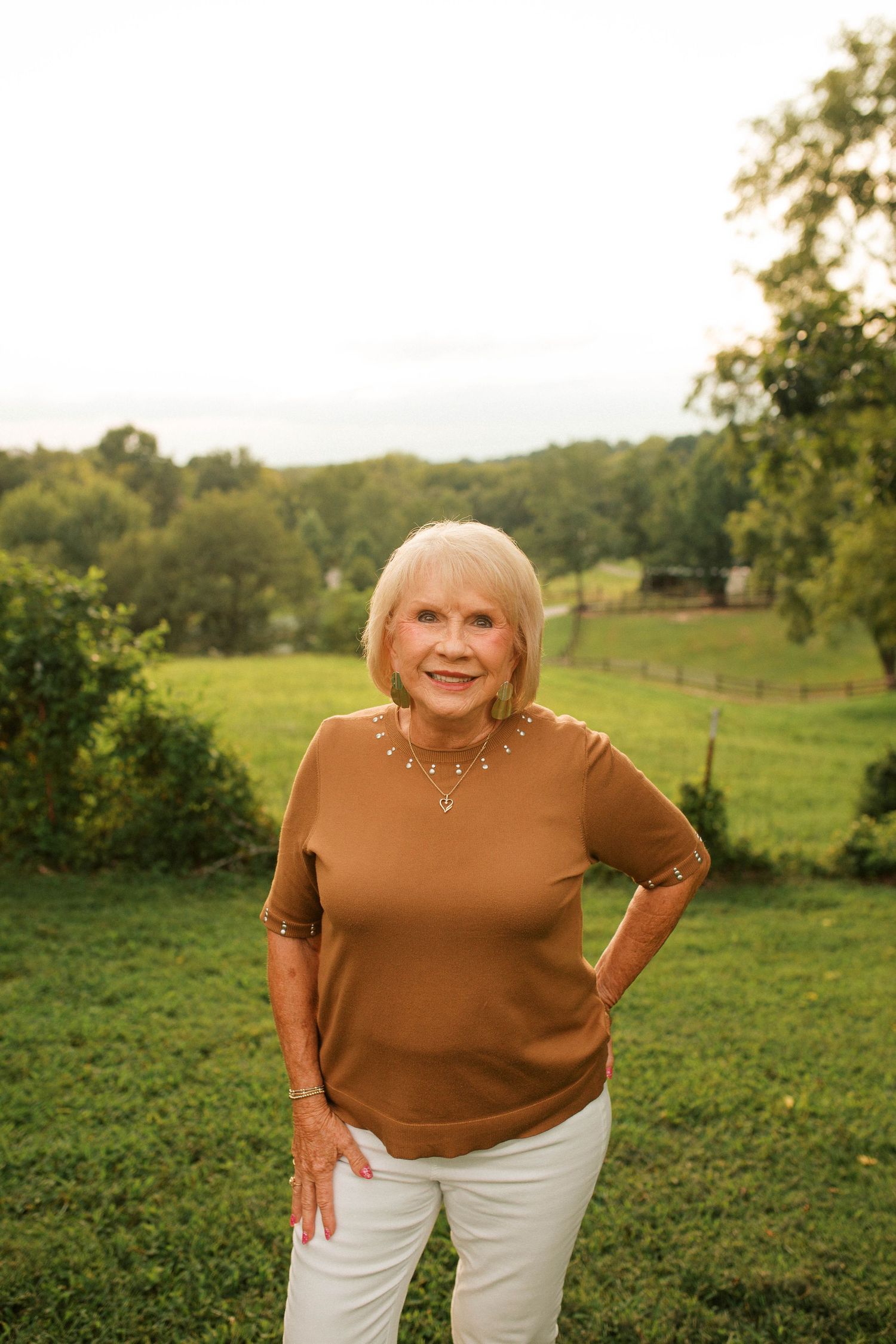 Smiling senior woman in brown top and white pants standing outdoors in lush green field with trees.