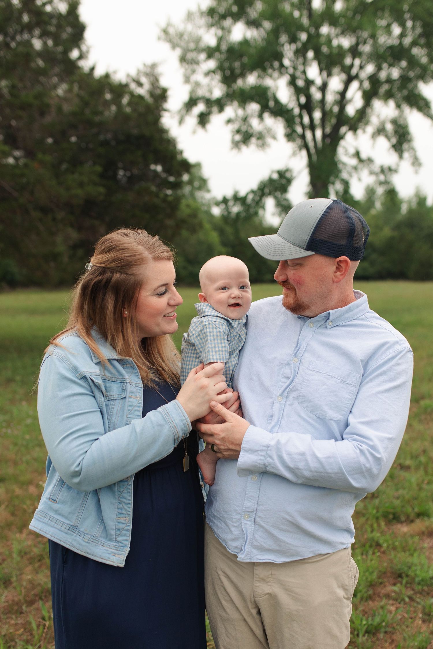 A young family smiles outdoors, with parents holding their laughing baby in a green field surrounded by trees.