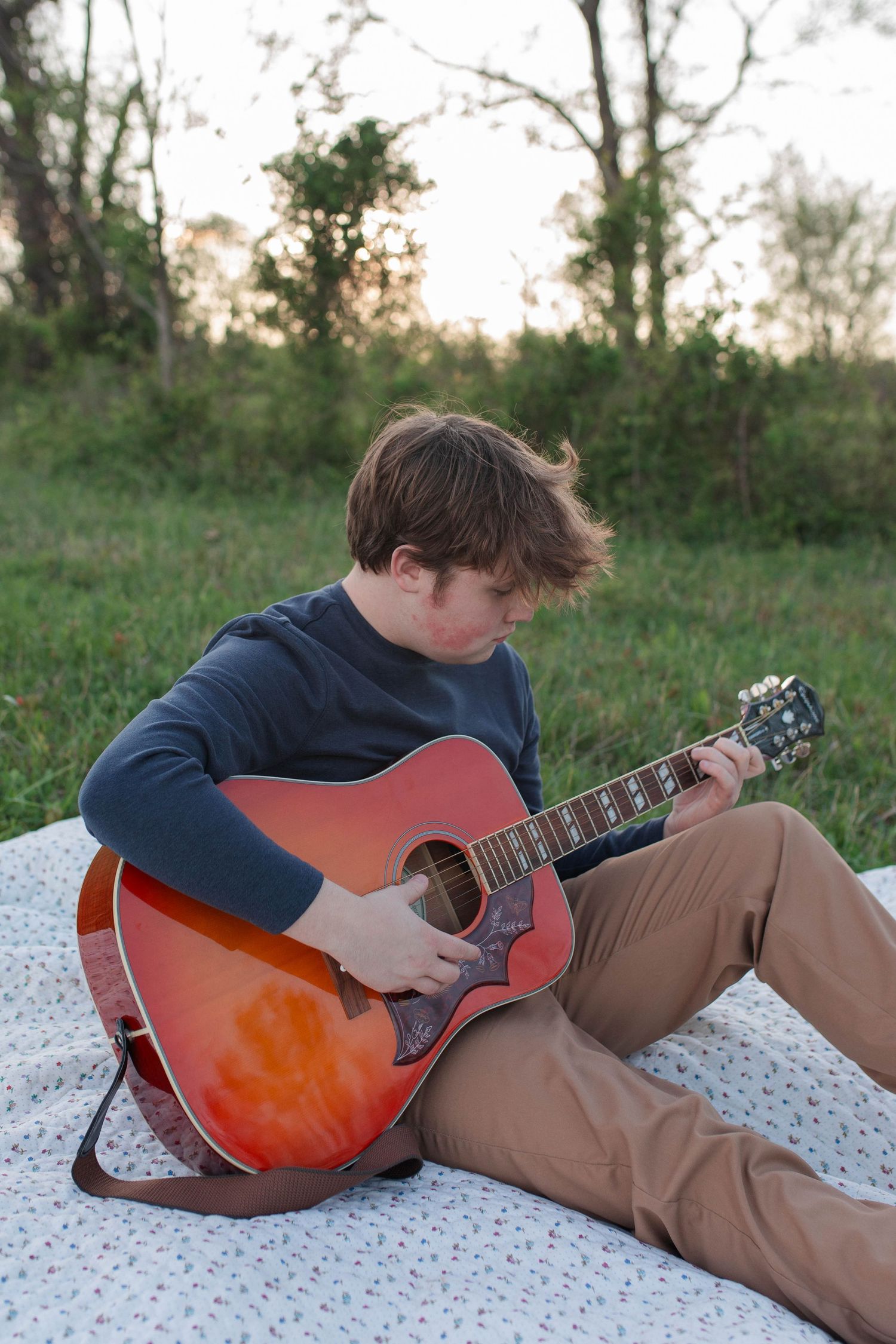 A young man sits on a blanket outdoors playing an acoustic guitar surrounded by green trees at sunset.
