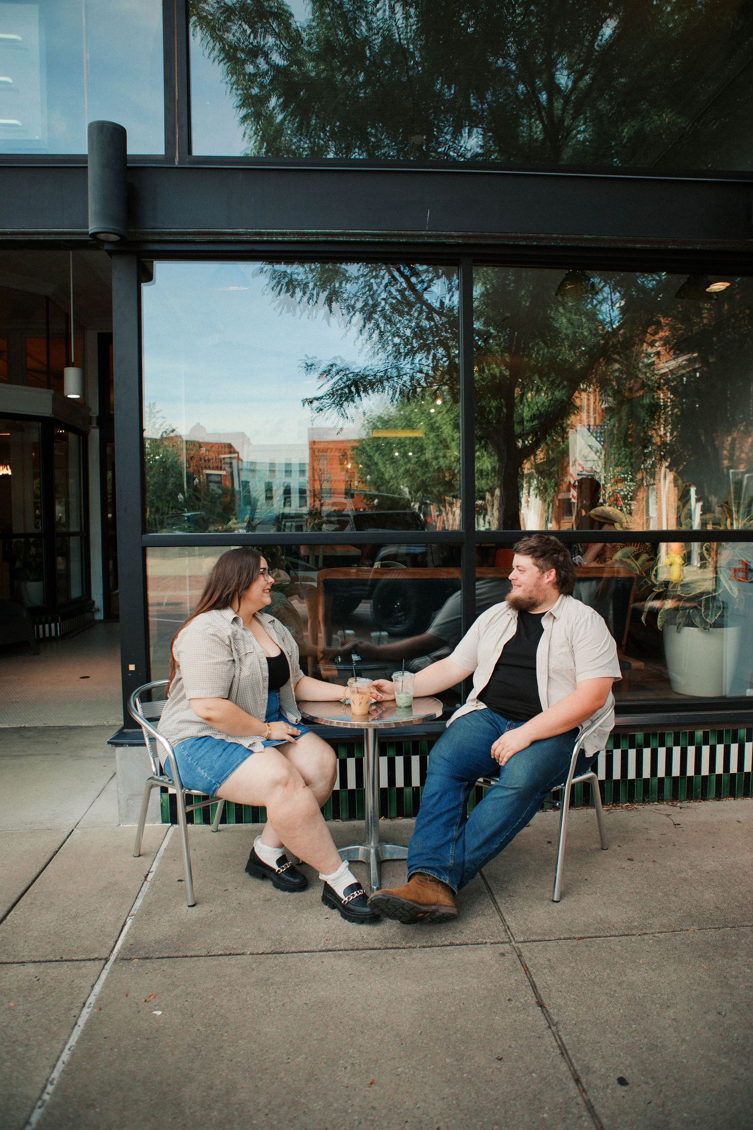 Couple holding hands at outdoor cafe table beside large glass windows on a sunny day.