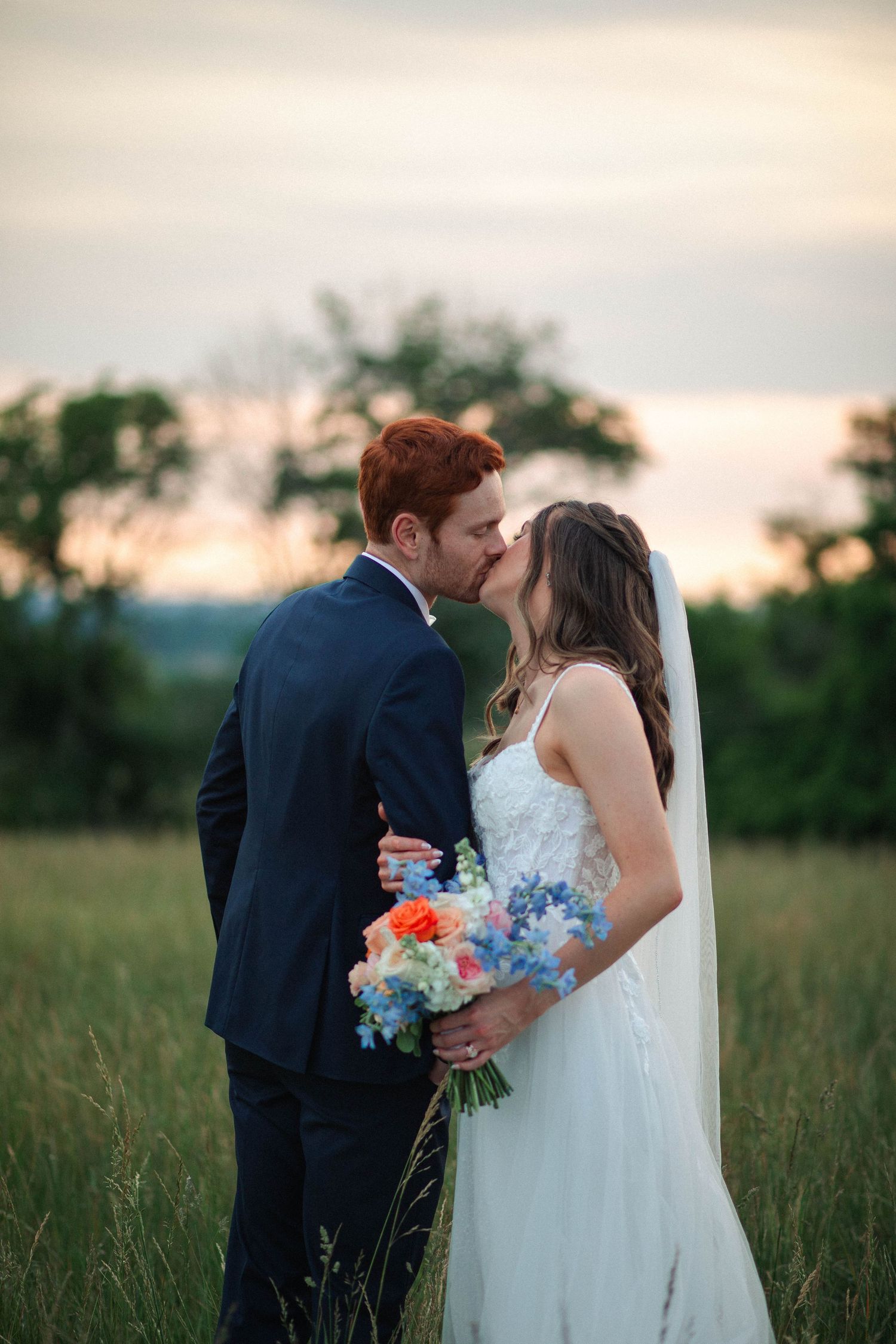 A bride and groom share a romantic kiss in a green field at sunset, bride holding a colorful bouquet of orange and blue flowers.