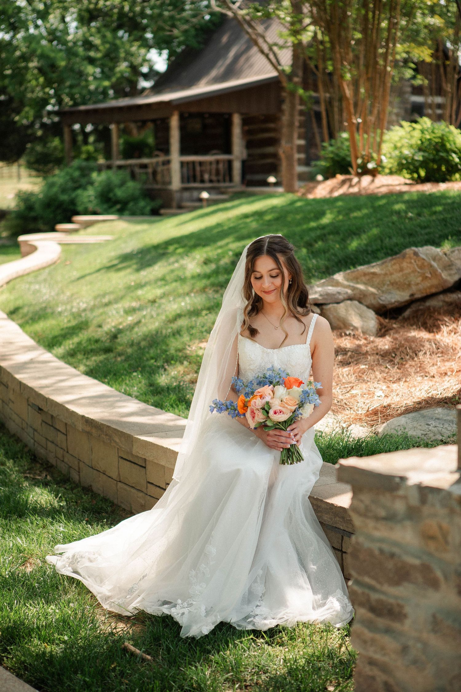 Bride in white gown and veil holds colorful bouquet of peach and blue flowers outdoors near rustic log cabin.