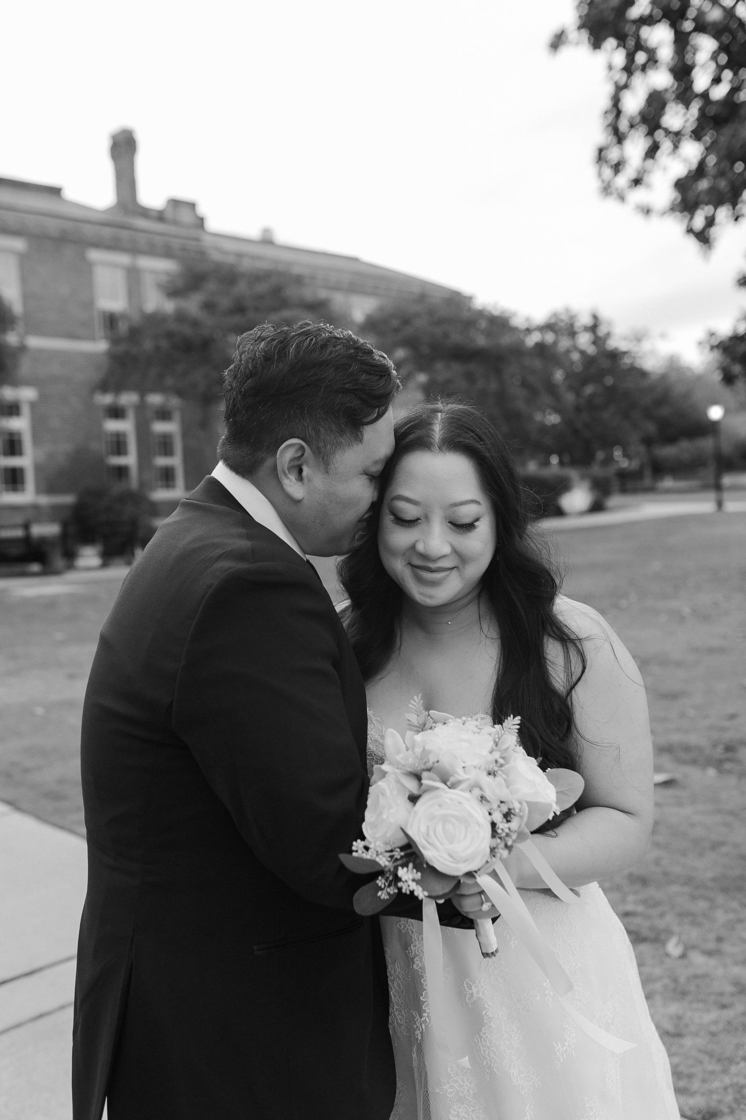 Black and white photo of a groom kissing bride on cheek as she smiles holding a bouquet.