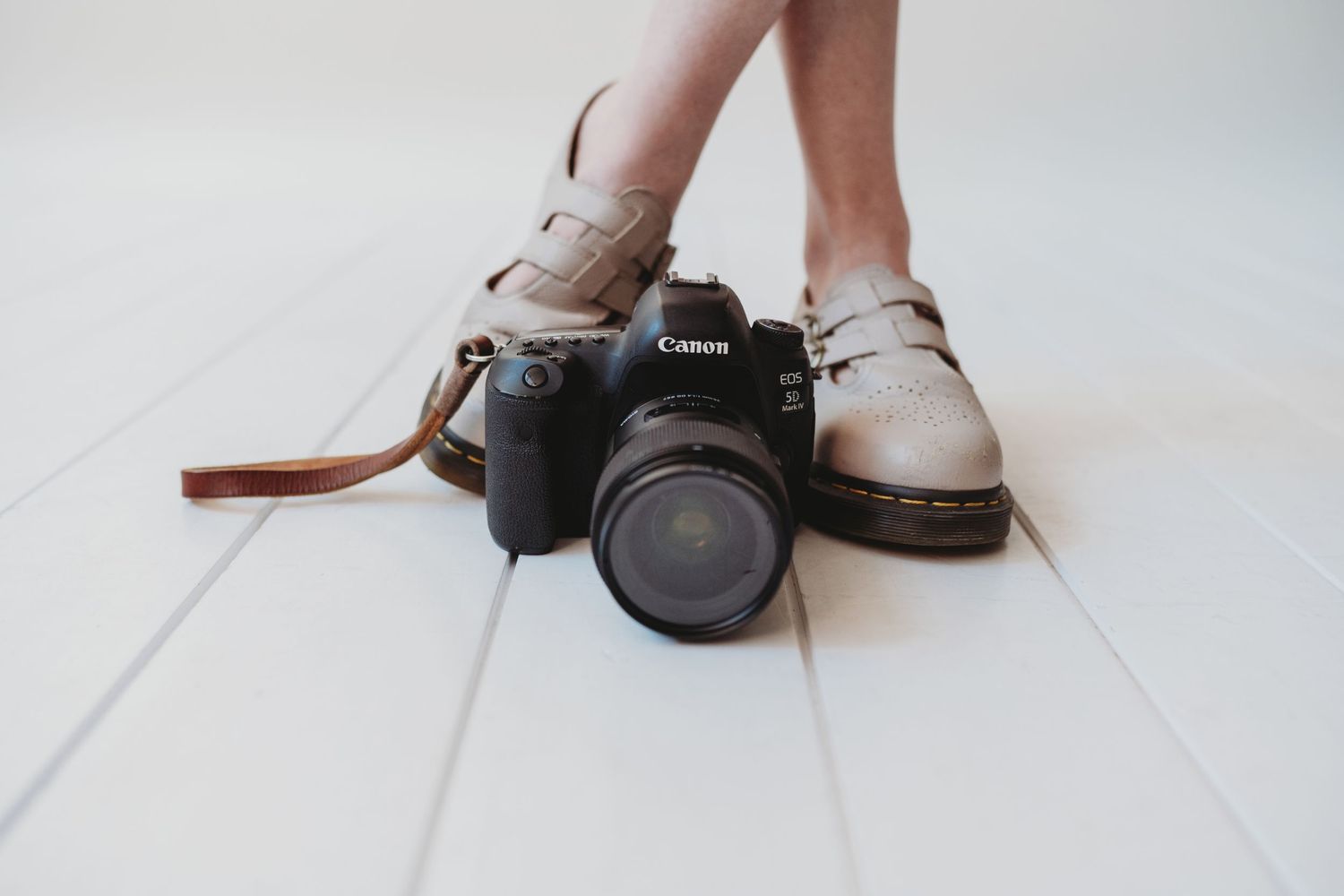 Canon DSLR camera on white wooden floor next to womens feet wearing beige leather shoes.