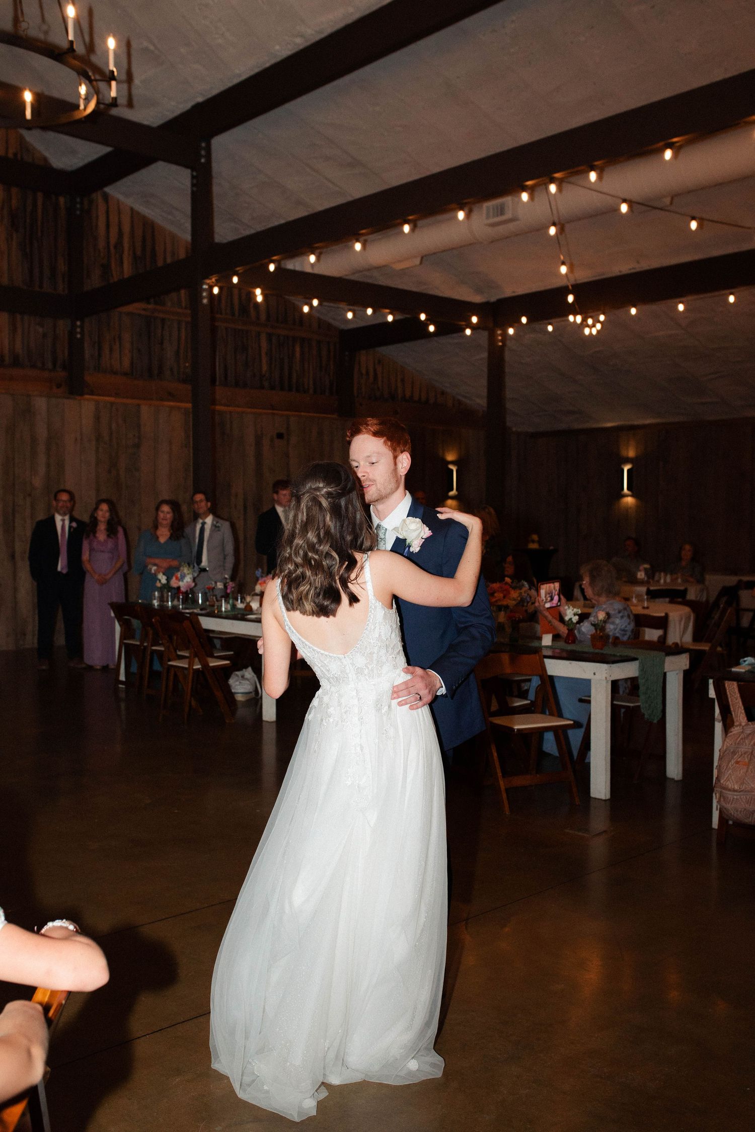Bride and groom share their first dance in a rustic barn venue adorned with string lights and surrounded by wedding guests.