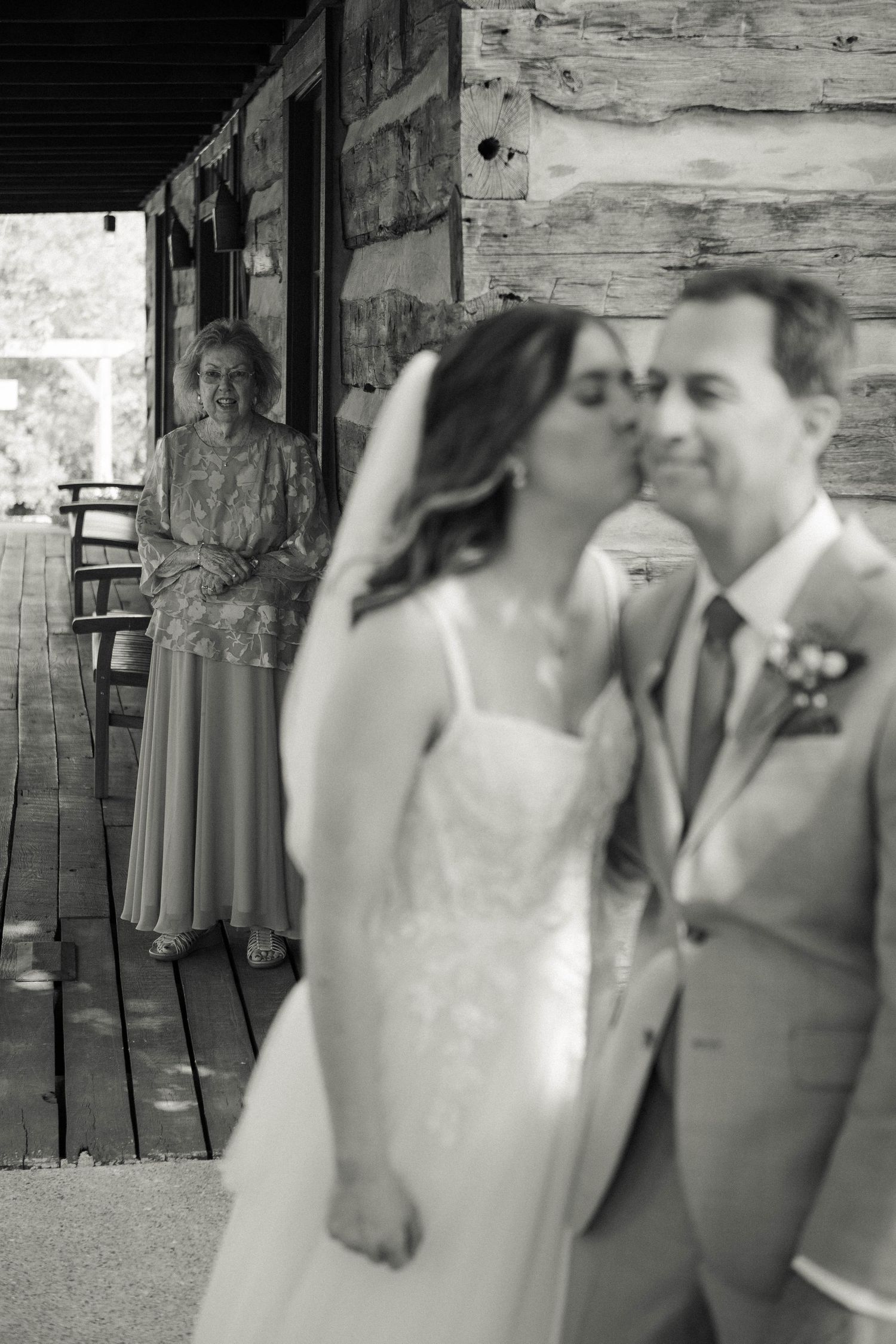 Black and white wedding photo of bride kissing groom while smiling woman watches from rustic wooden porch backdrop.