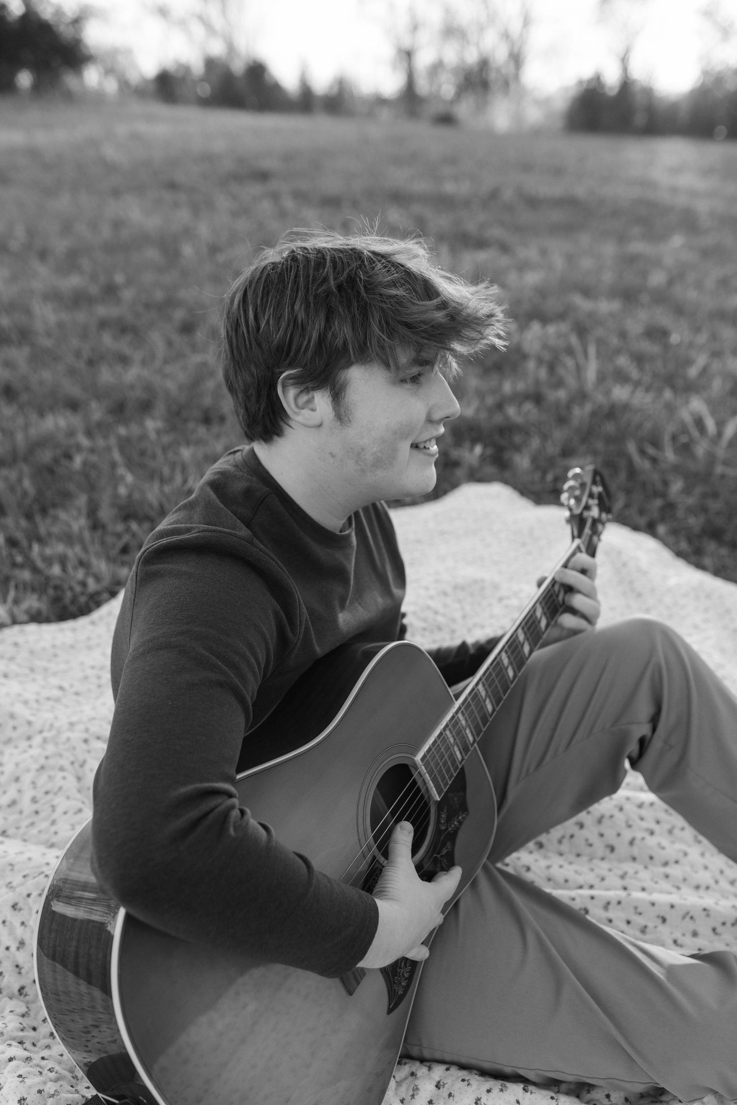 Young man playing acoustic guitar outdoors on a blanket in a grassy field, black and white photo.