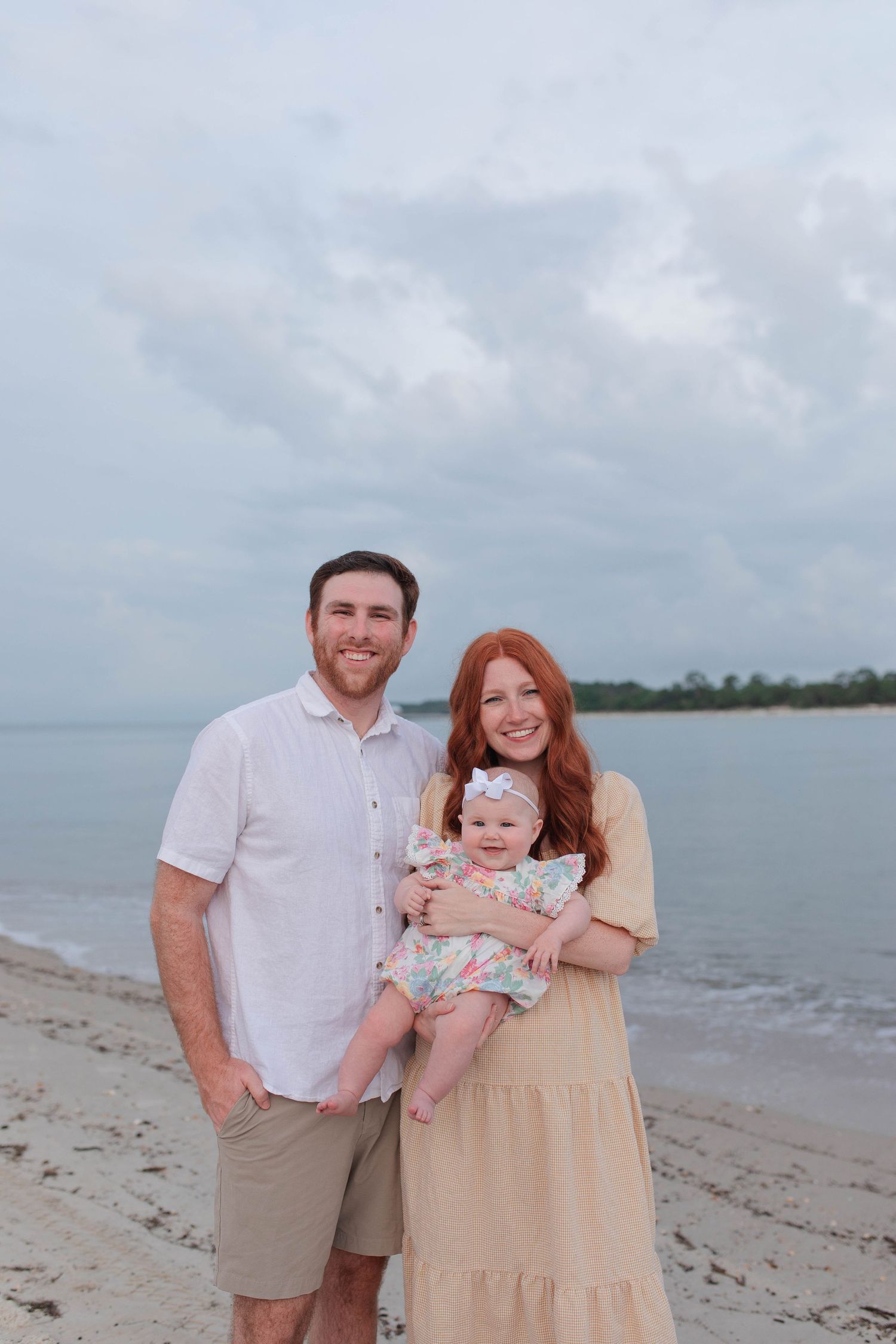 Young family with red-haired mother, father, and baby posing on a sandy beach with cloudy skies.