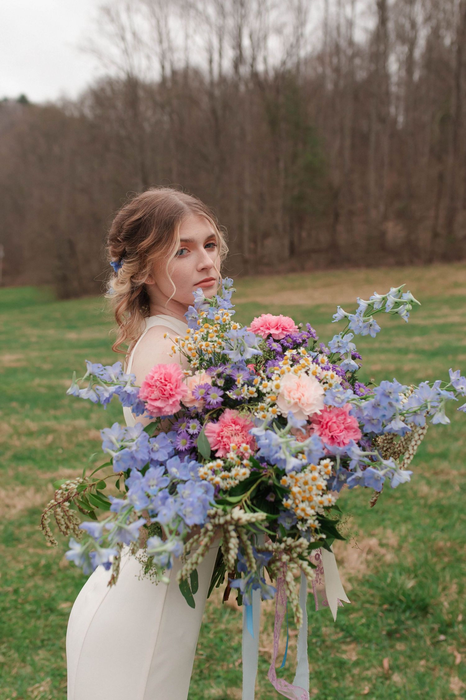 Bride holds an oversized colorful bouquet of pink and blue flowers in a green field wearing a white gown after eloping in Nashville.