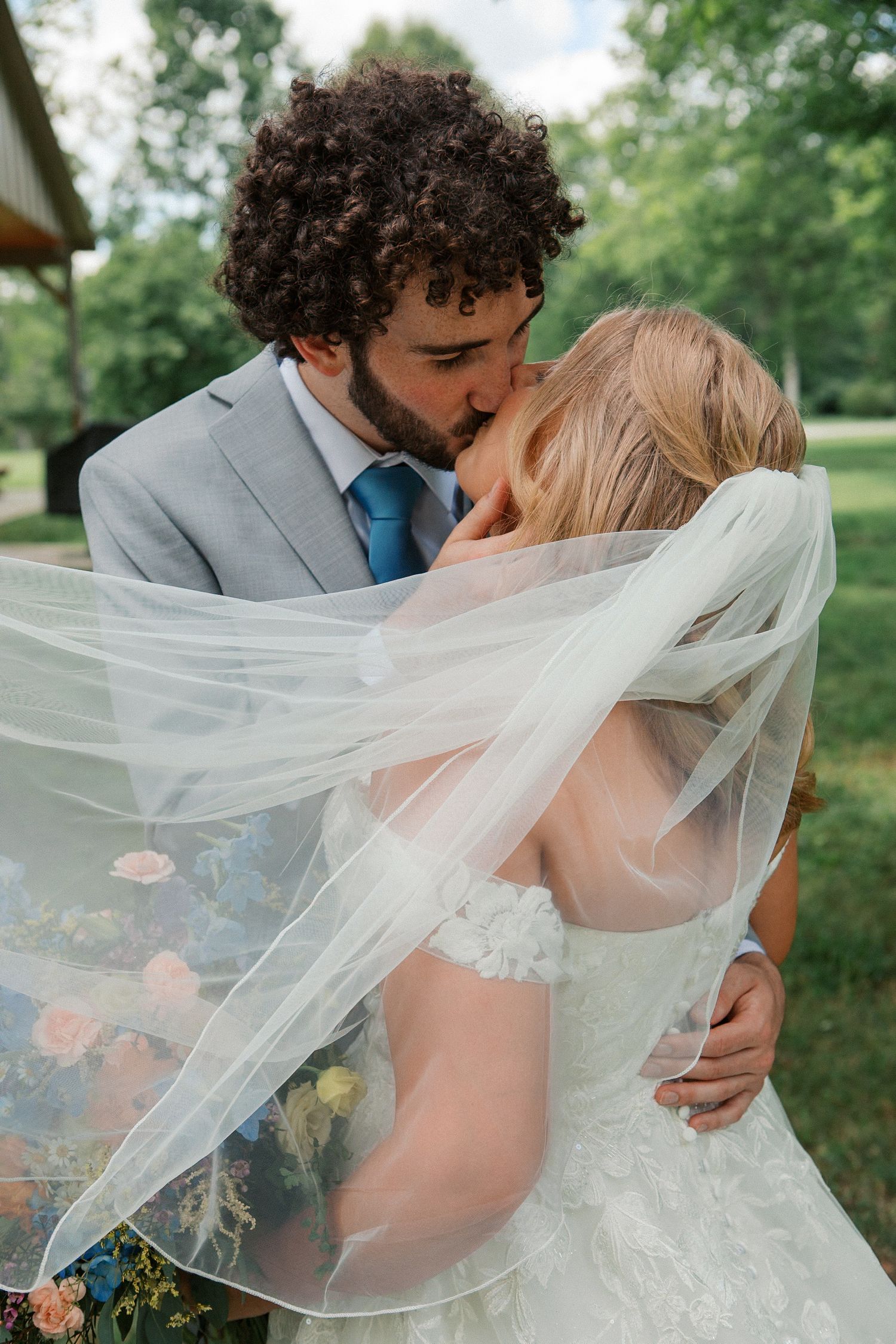 A bride and groom share a romantic kiss outdoors, with her flowing veil billowing in the breeze as she holds a colorful bouquet.