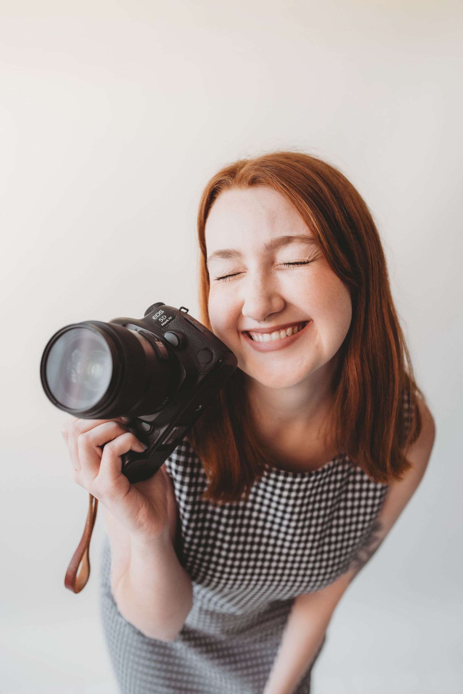 Smiling redhead woman holding a DSLR camera with eyes closed, wearing a checkered dress against a white background.