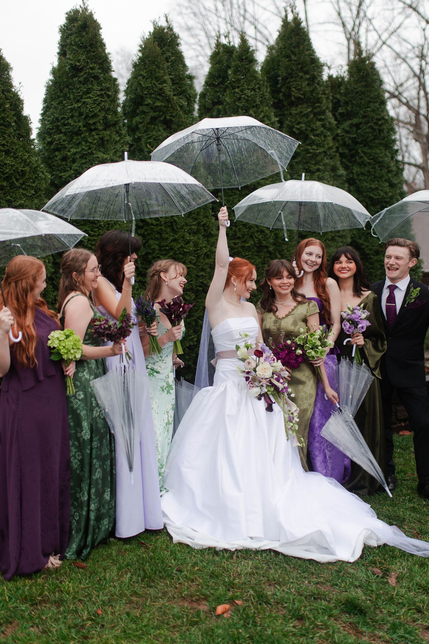 Bride raises clear umbrella celebrating with wedding party in mismatched green and purple dresses outdoors after micro wedding in Nashville.