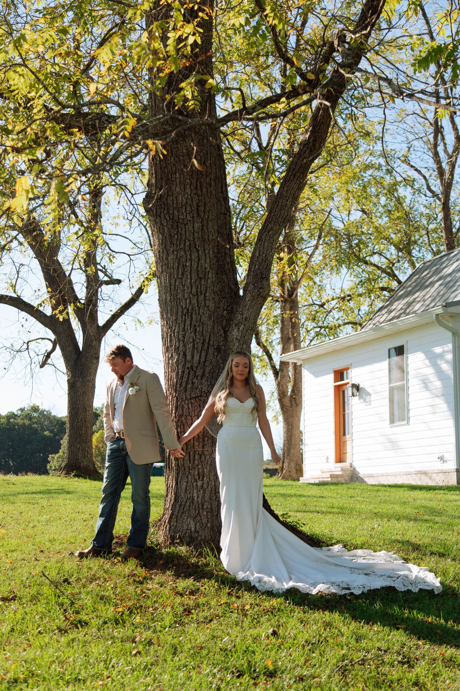 Bride in white gown and groom in tan jacket hold hands by a large tree on a sunny day near a white farmhouse.