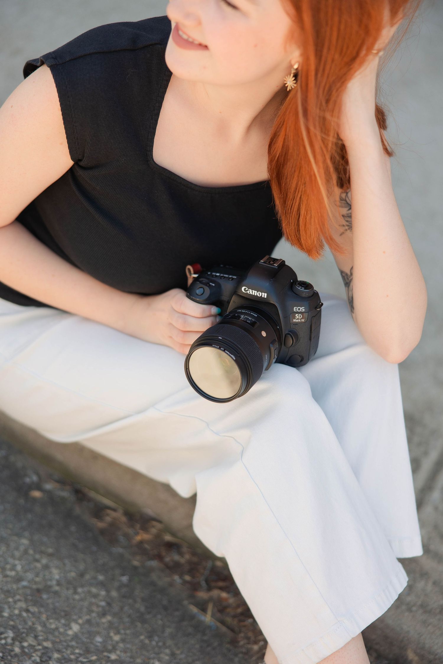 Red-haired woman in black top and white pants sitting outdoors holding a Canon EOS DSLR camera.