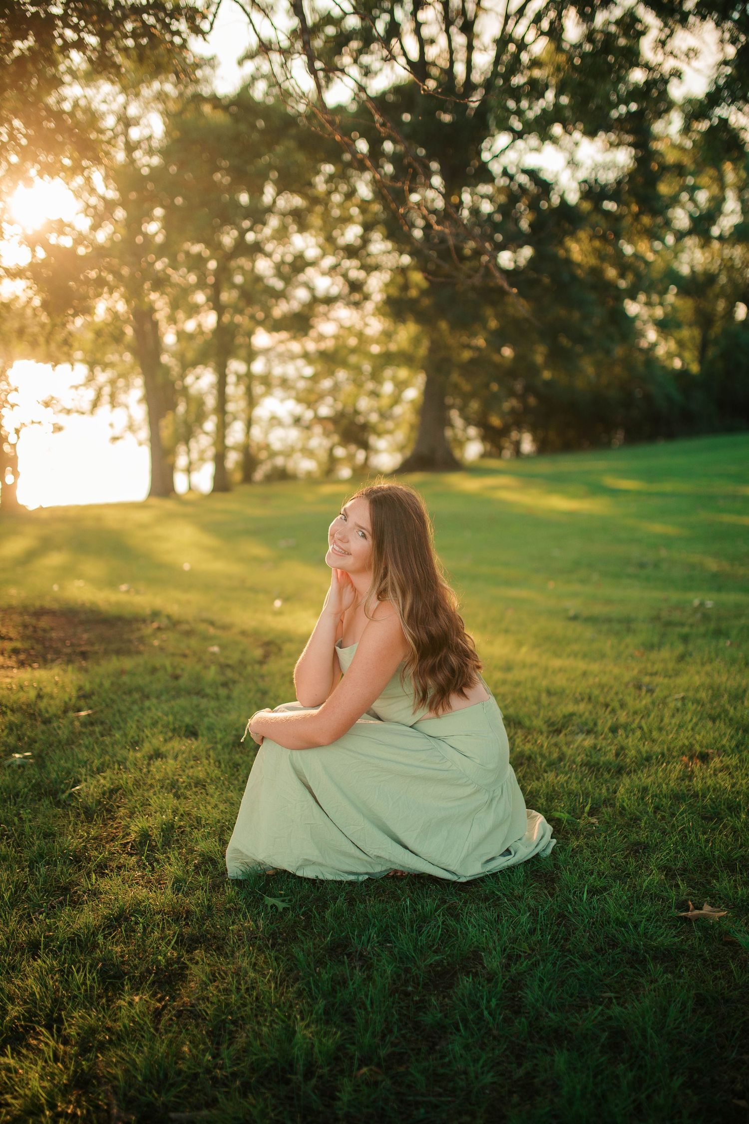 Young woman in mint green dress sitting gracefully on grass during golden hour sunset in park.