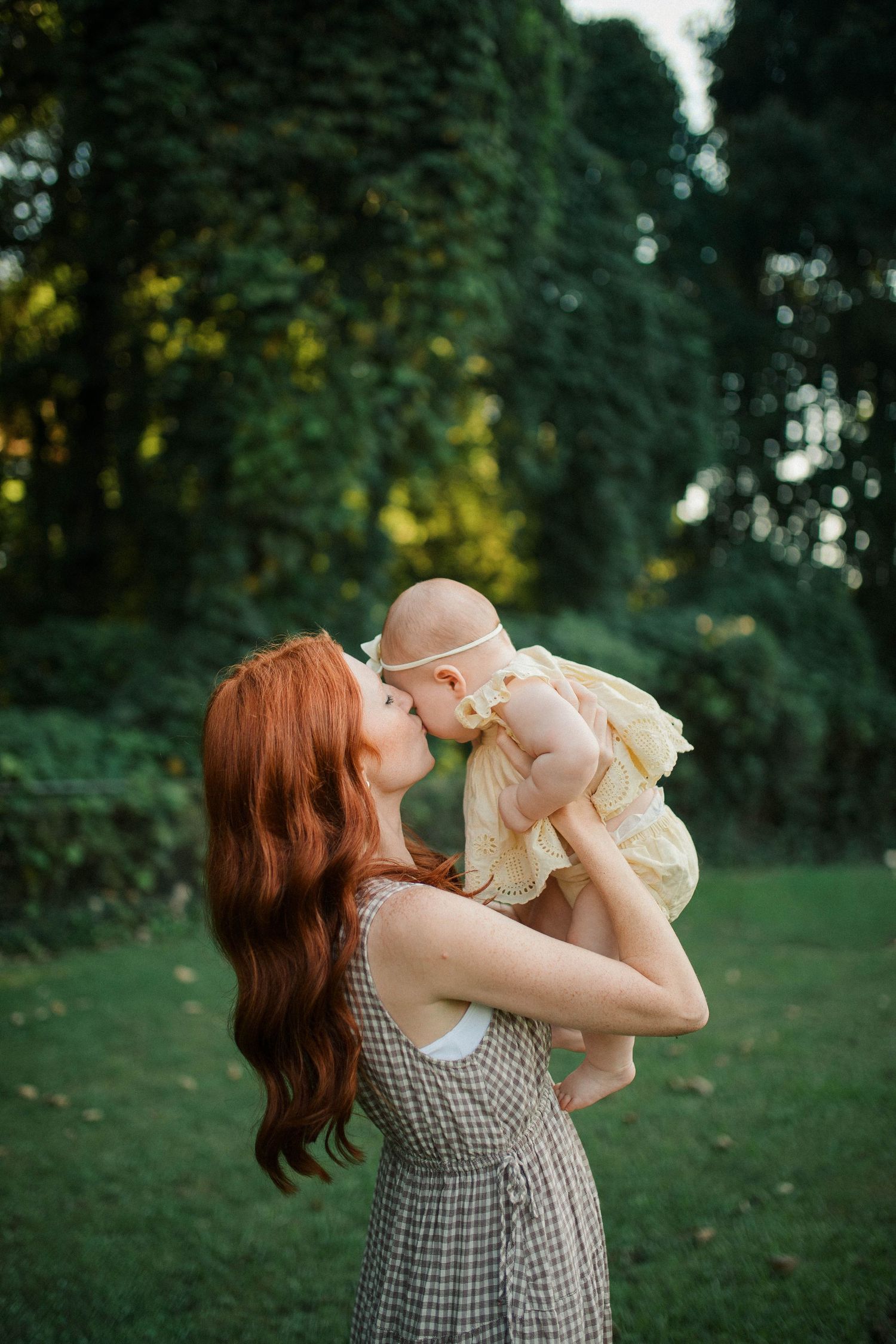 A red-haired mother in a plaid dress kisses her baby girl in a yellow dress outdoors in a lush green park.