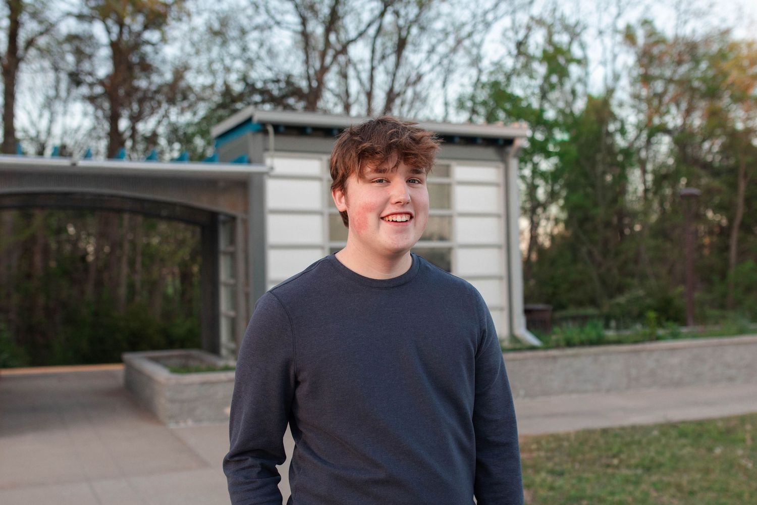 Smiling young man in navy blue sweatshirt standing outdoors near a modern structure surrounded by trees.