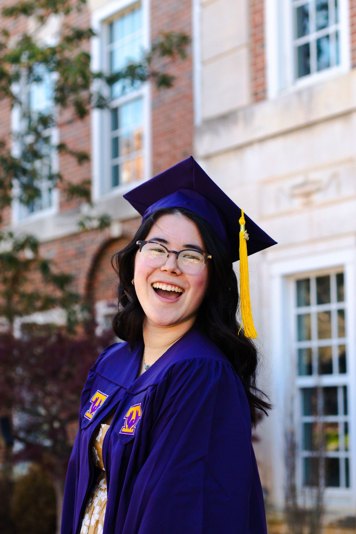 Smiling young woman in purple graduation cap and gown with gold tassel poses outdoors in front of brick building.