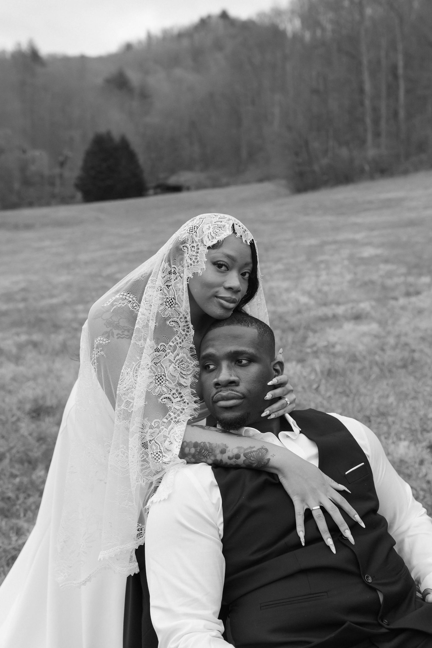 Black and white portrait of bride and groom sitting in a field, bride wearing lace veil embracing groom after eloping in Nashville.
