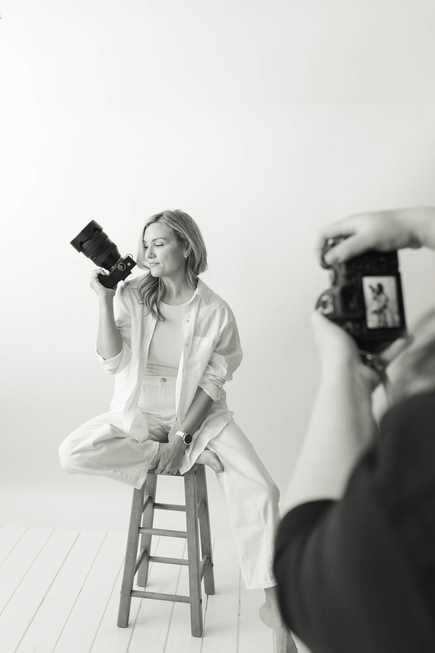 Black and white photo of a woman in white outfit holding a camera on a stool while being photographed in a studio.