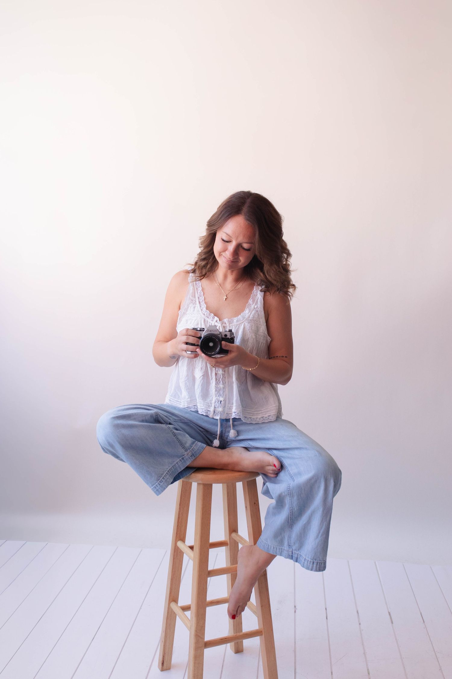Woman sitting on wooden stool holding vintage film camera, wearing white top and blue jeans, smiling in bright studio.