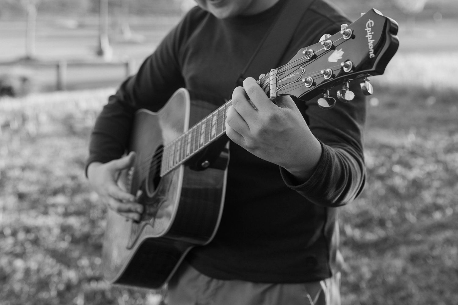 Person playing an acoustic guitar outdoors in a black and white photo, strumming chords with focused hands.