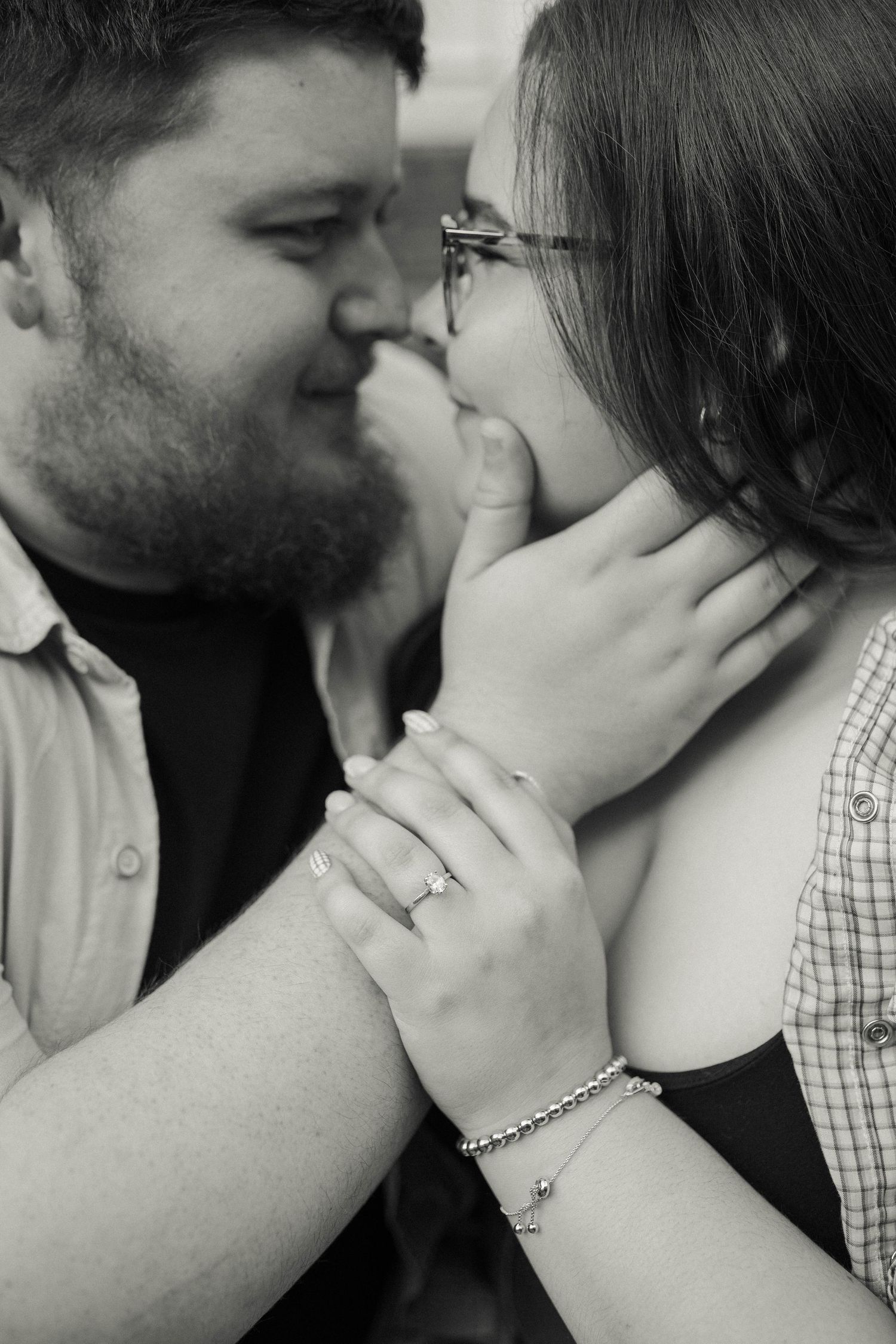 Black and white photo of a couple about to kiss, woman wearing engagement ring and bracelet, man tenderly holding her face.