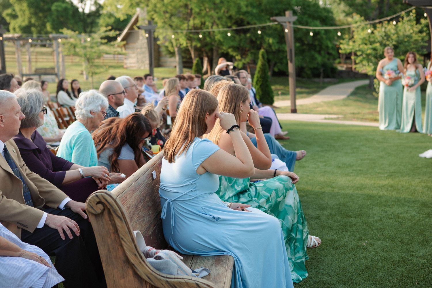 Guests seated on wooden benches at an outdoor wedding ceremony on a green lawn with string lights.