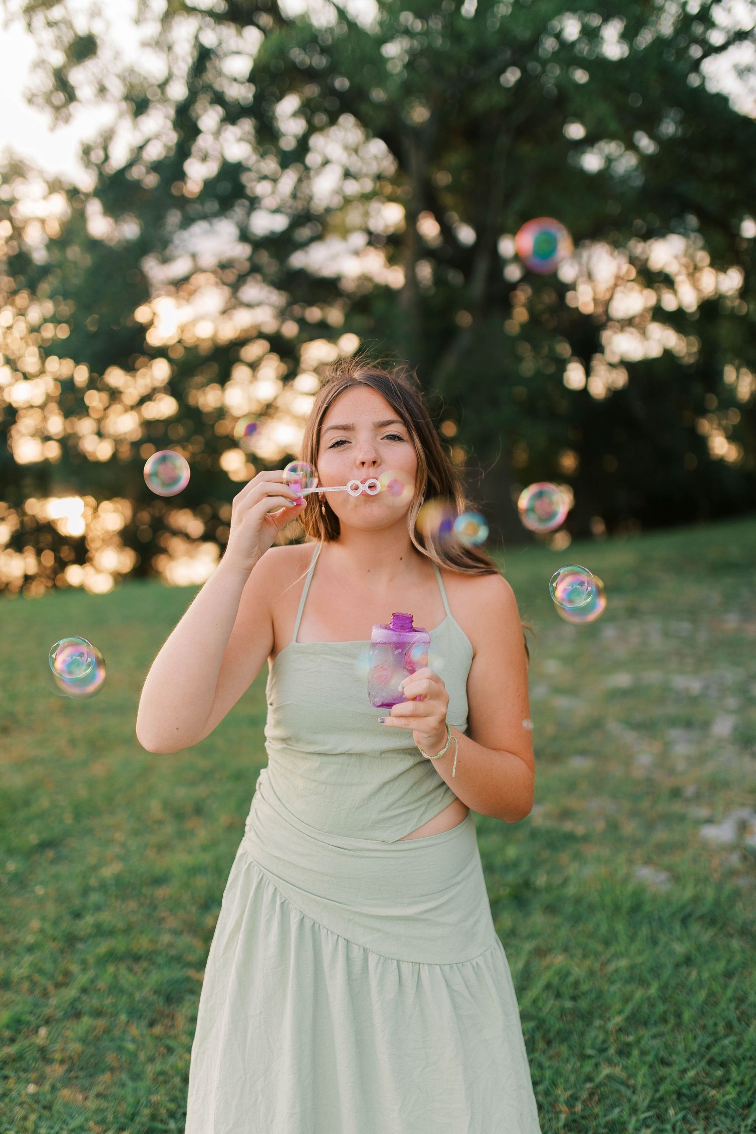Young woman in mint green dress blowing colorful soap bubbles outdoors in a sunlit park setting.