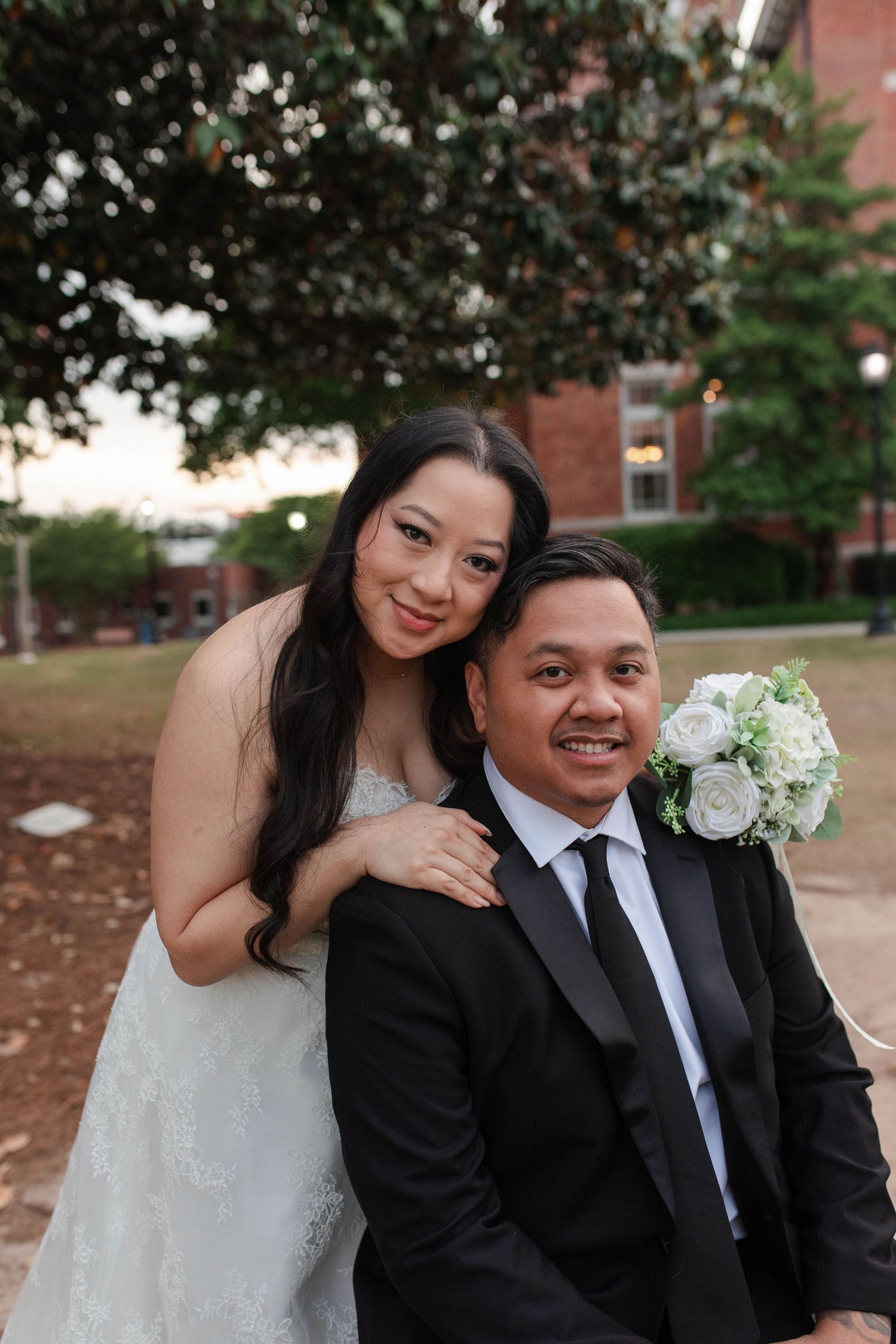 A smiling bride and groom pose outdoors in wedding attire, she holds white roses bouquet, brick building in background.