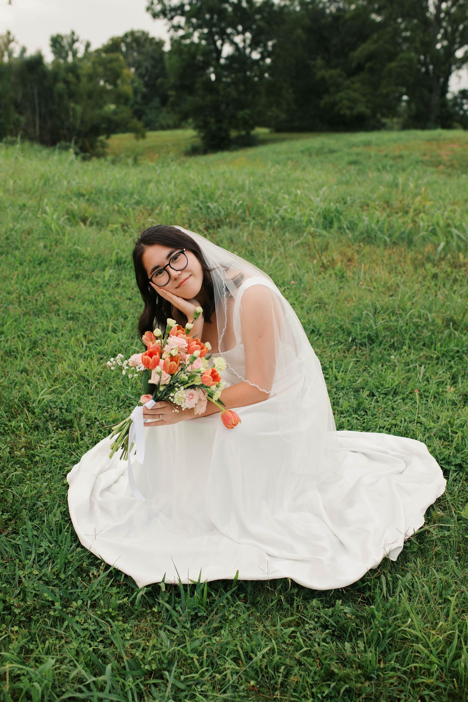 Bride in white gown and veil sits on green grass holding colorful orange and pink bouquet, smiling at camera.