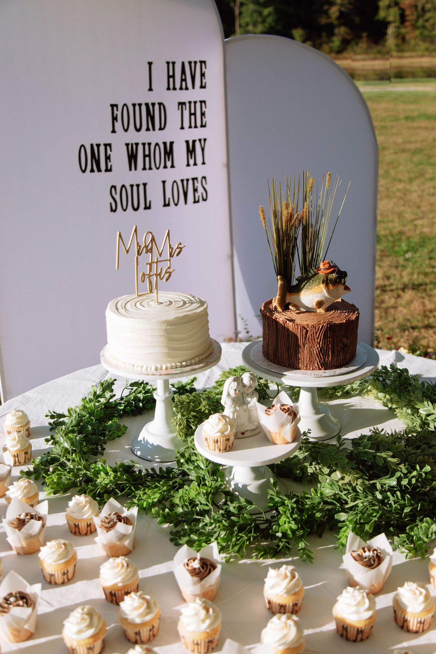 Rustic wedding dessert table with white cake, fish-topped groom's cake, cupcakes, and greenery under a quote sign.