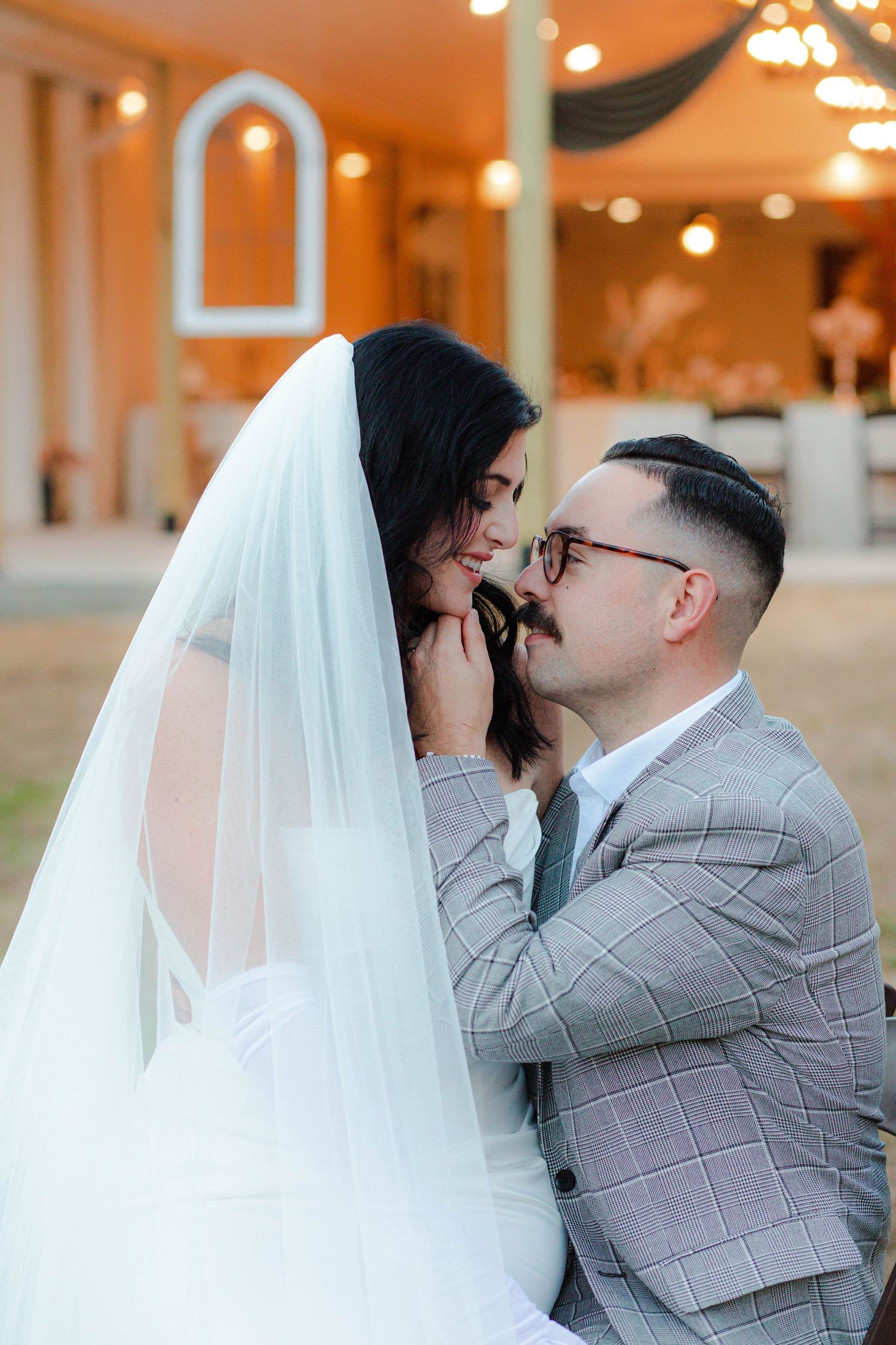 Groom in plaid suit tenderly holds bride's face under her veil in a warmly lit outdoor wedding venue after  micro wedding in Nashville.