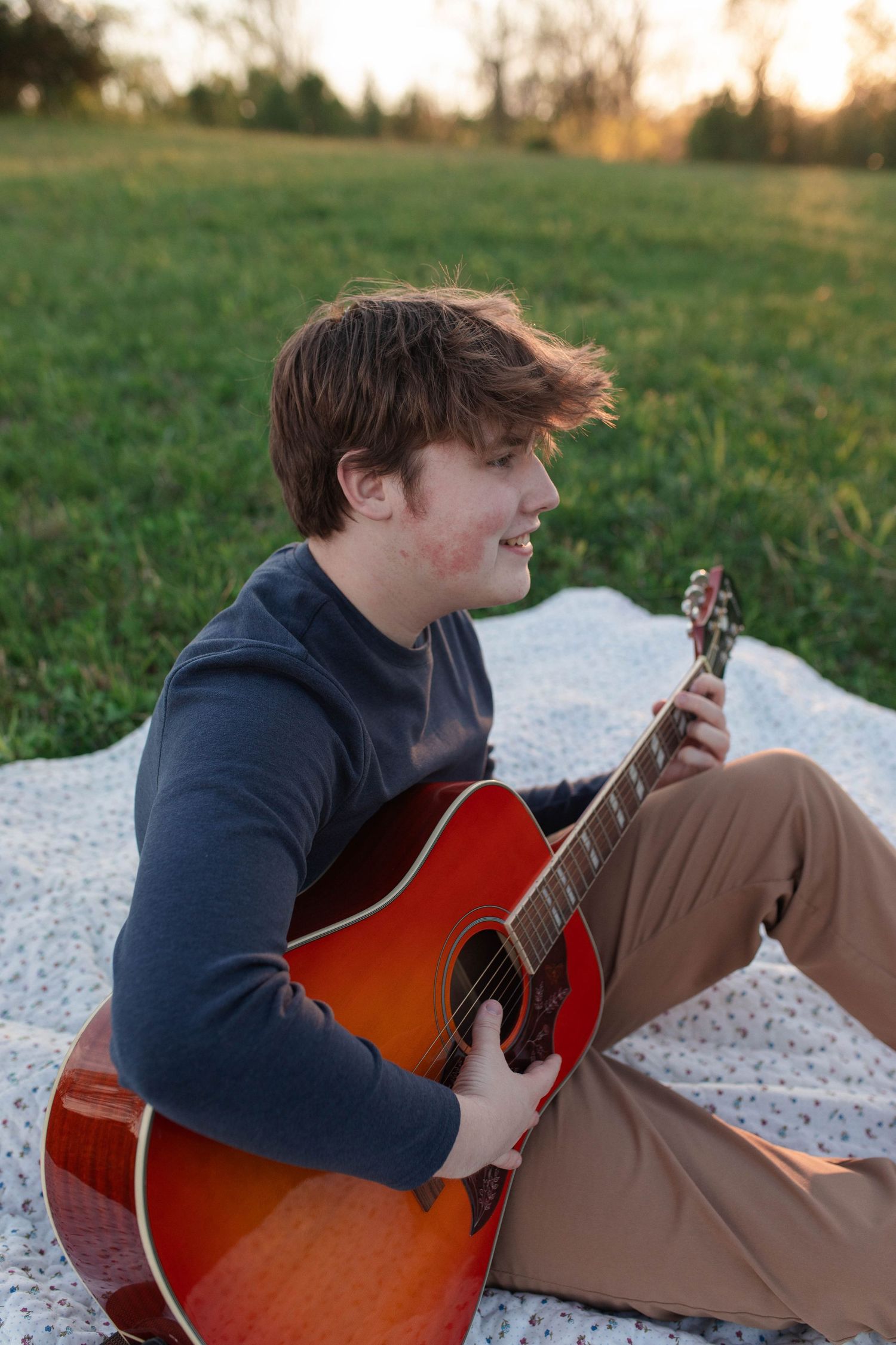 Young man playing acoustic guitar on a blanket in a green field at sunset, smiling outdoors.