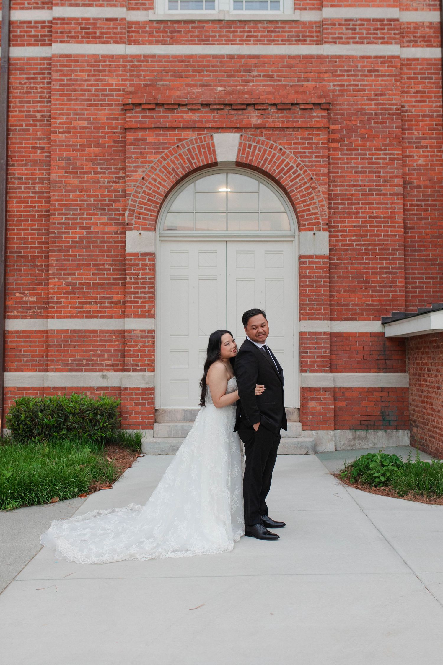 Bride and groom posing together in front of a red brick church with arched white doors on their wedding day.