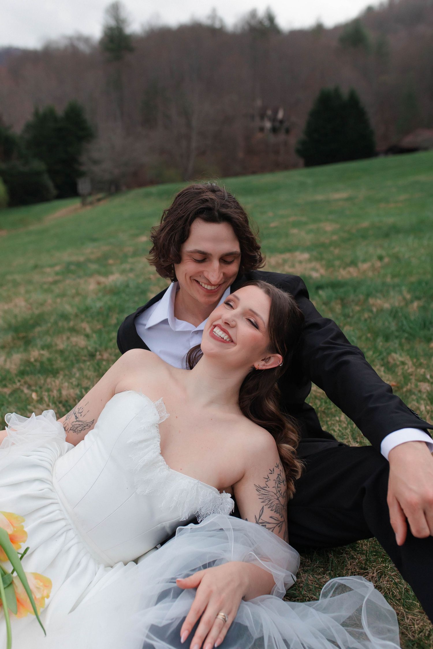 Bride and groom sitting on green grass, smiling and embracing outdoors with wooded hills in background  after eloping in Nashville.