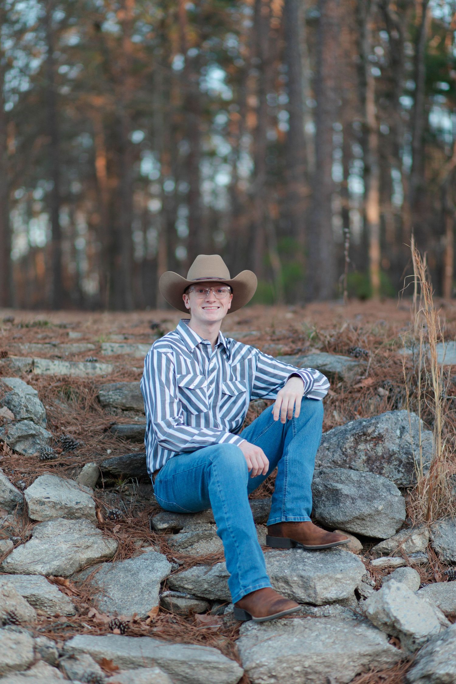 Young man in cowboy hat and striped shirt sitting on rocks in a pine forest in Nashville.
