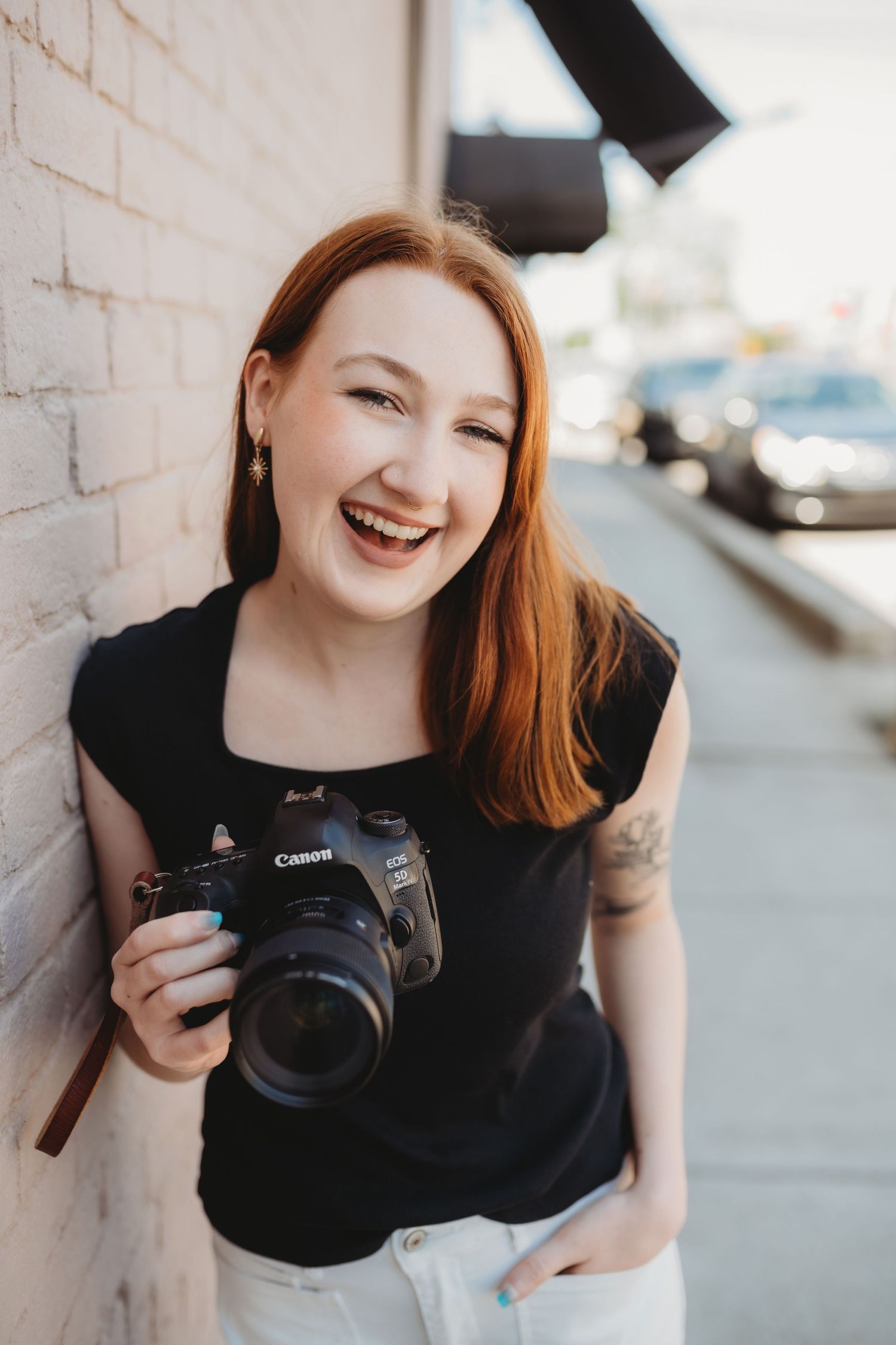 Smiling redhead woman holding a Canon DSLR camera, leaning against a brick wall outdoors in a black top.