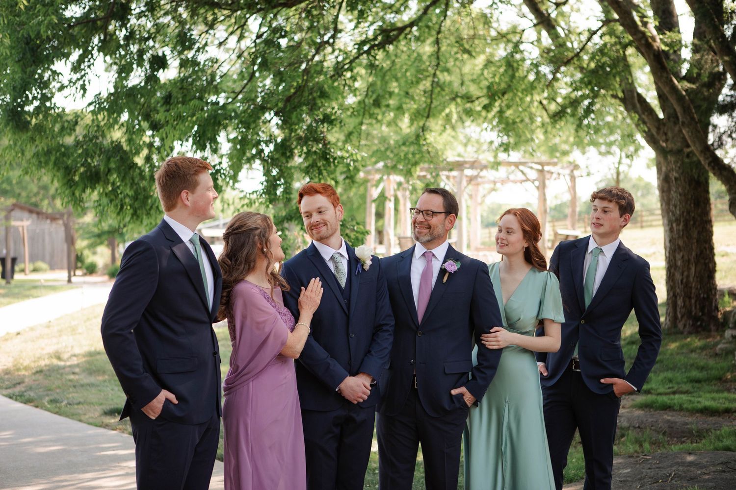 Wedding party poses outdoors under trees, groom in navy suit with pink tie, bridesmaids in purple and sage green dresses.