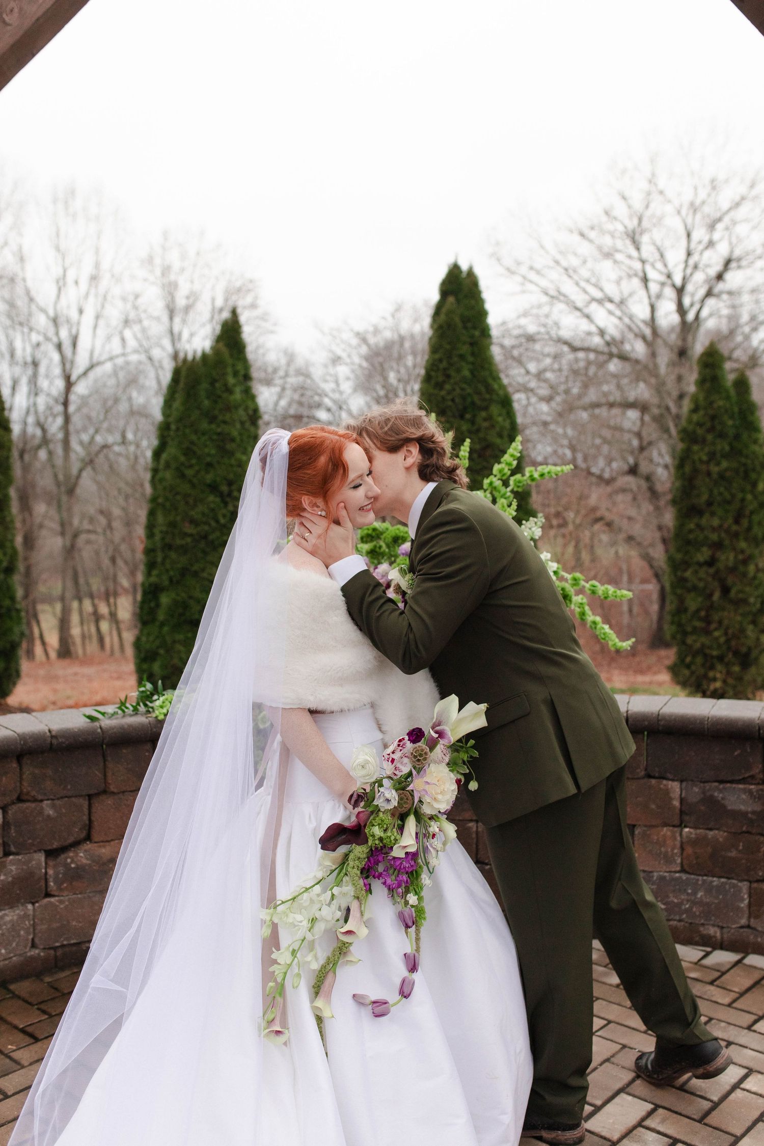 Red-haired bride in white gown with fur wrap kissing groom in olive suit under garden archway after micro wedding in Nashville.