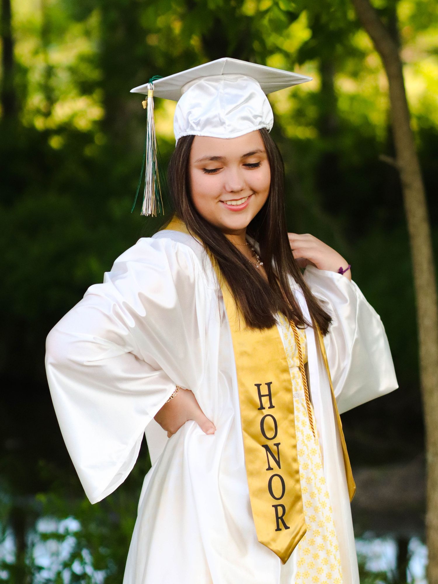 Smiling graduate in white cap and gown with gold Honor stole posing outdoors among green trees.